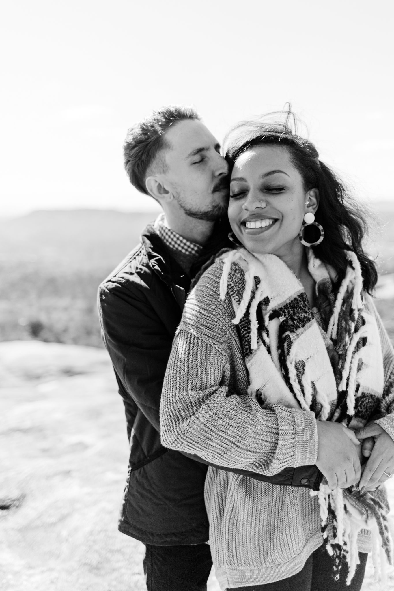 A romantic black and white portrait of a couple embracing outdoors with mountains in the background.