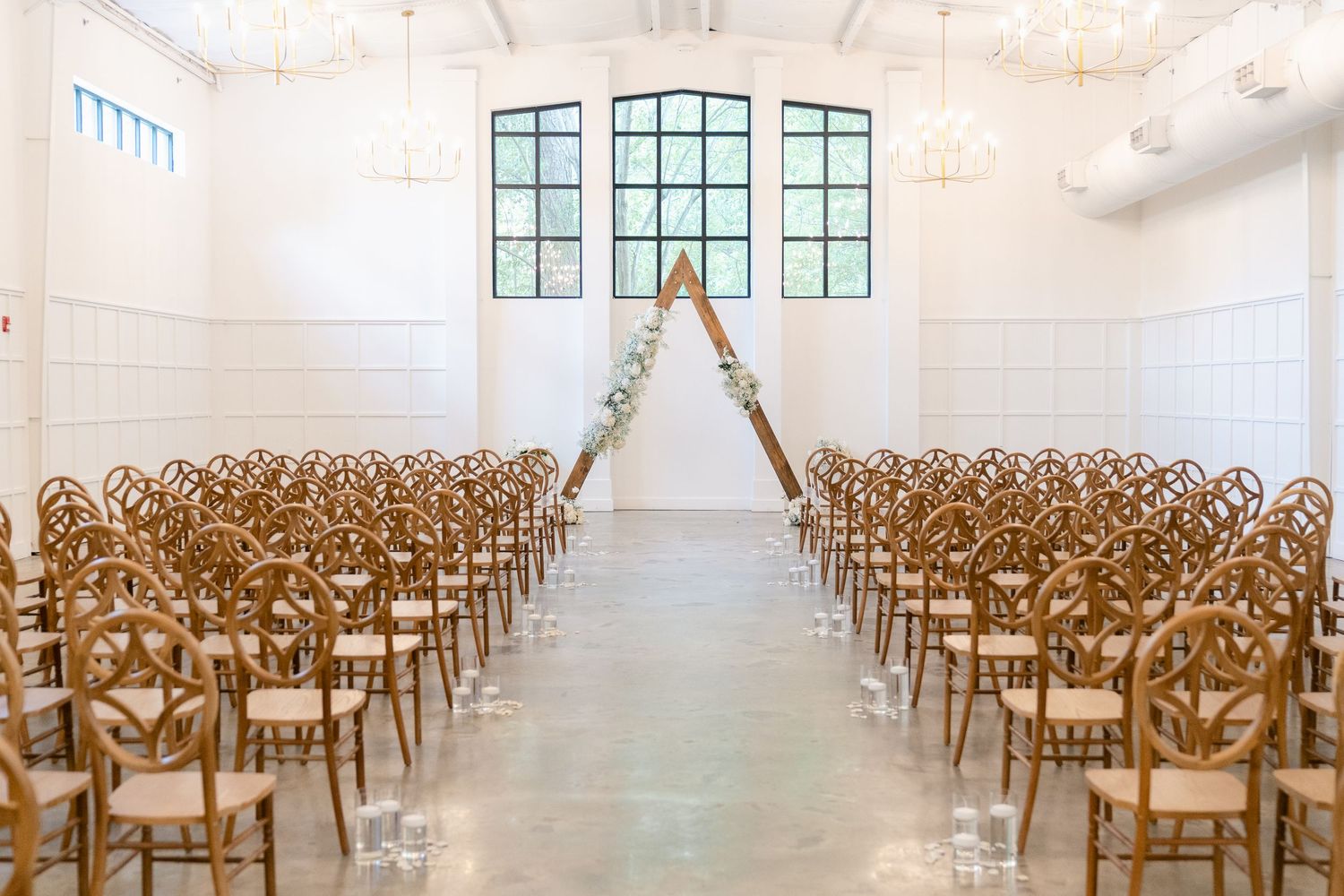 A bright white wedding venue with wooden chairs arranged in rows features tall windows and natural lighting.