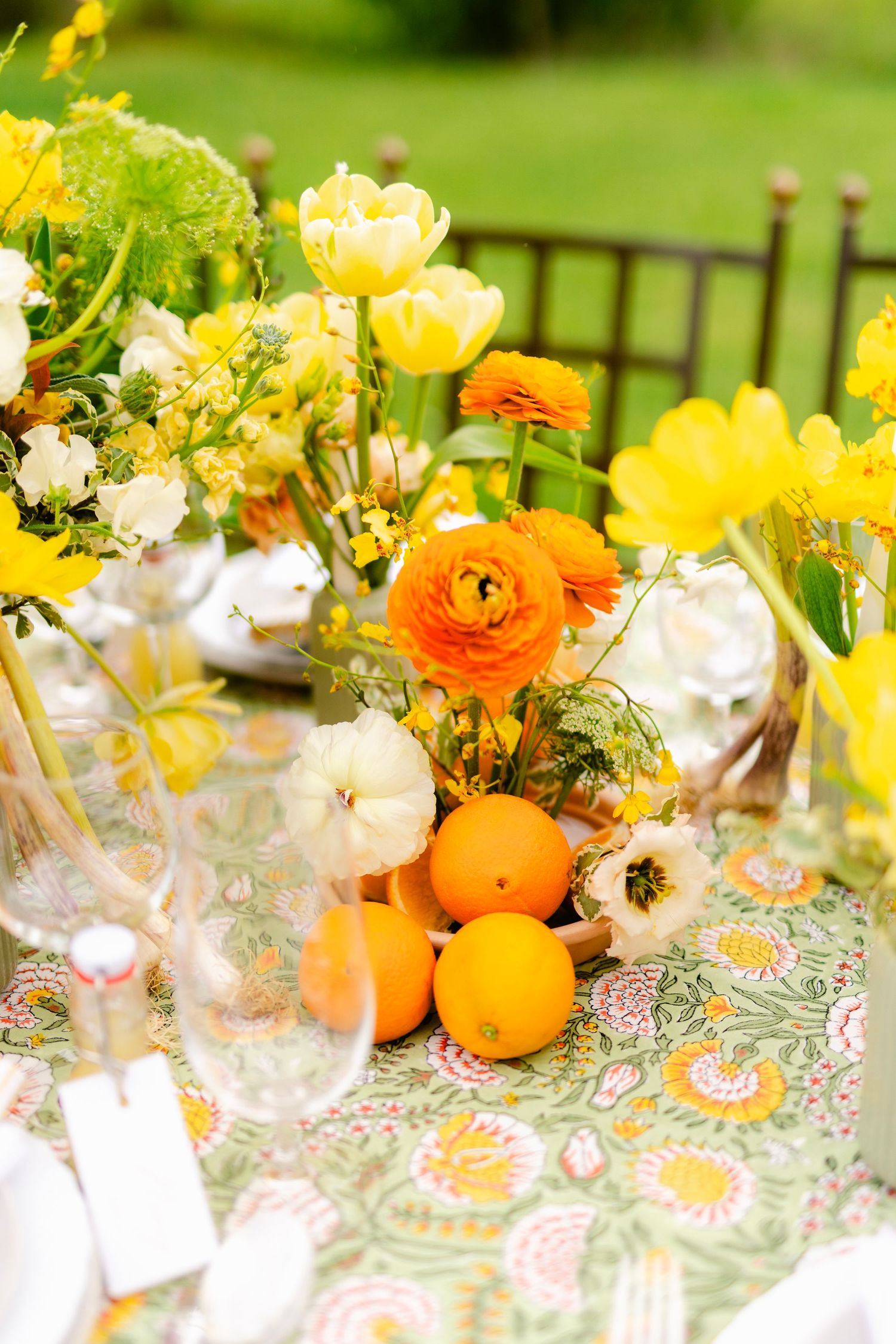 Elegant spring table setting with yellow ranunculus flowers, oranges, and delicate glassware on a floral tablecloth.