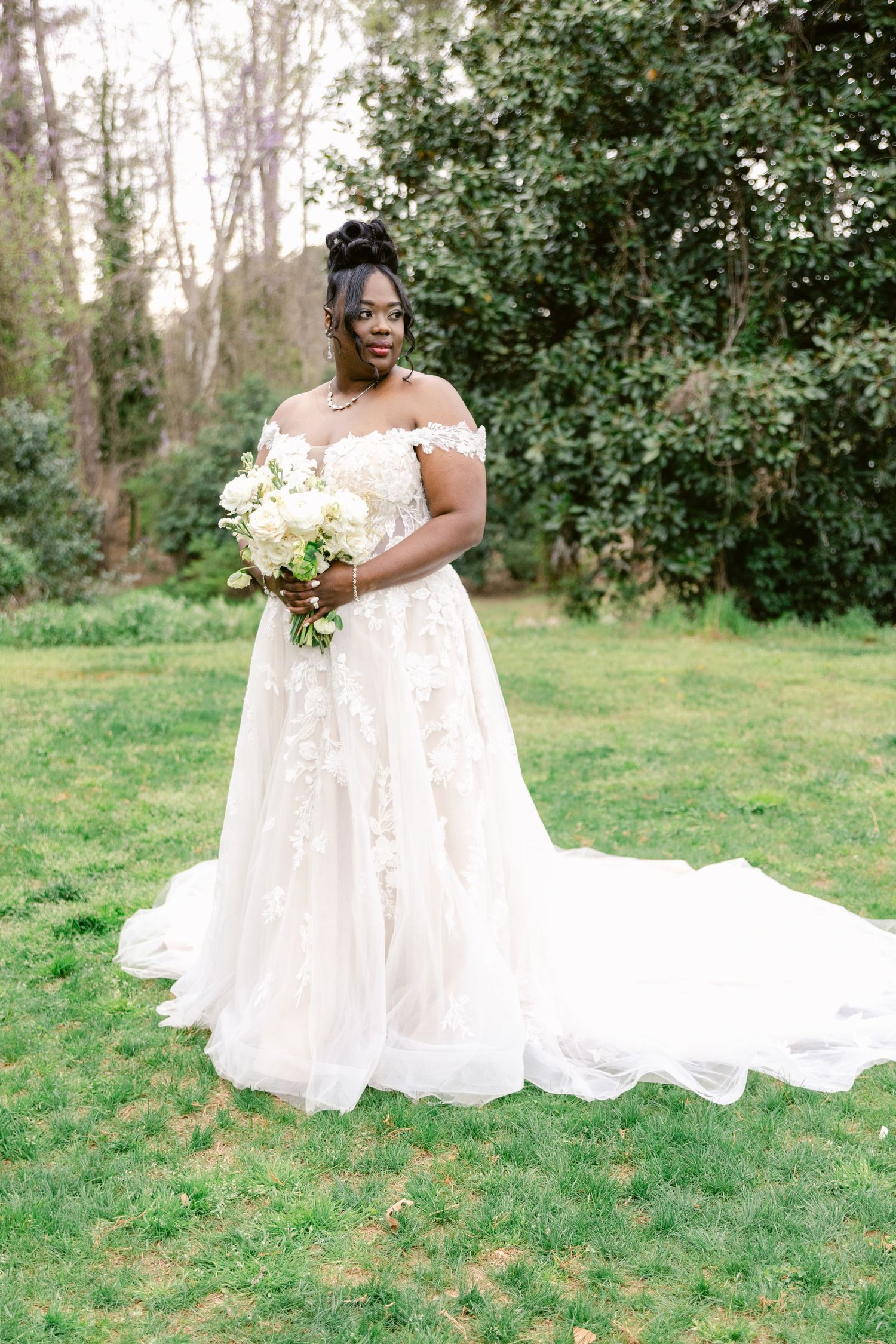 Elegant white lace wedding dress with flowing train cascades across green grass in outdoor garden setting.