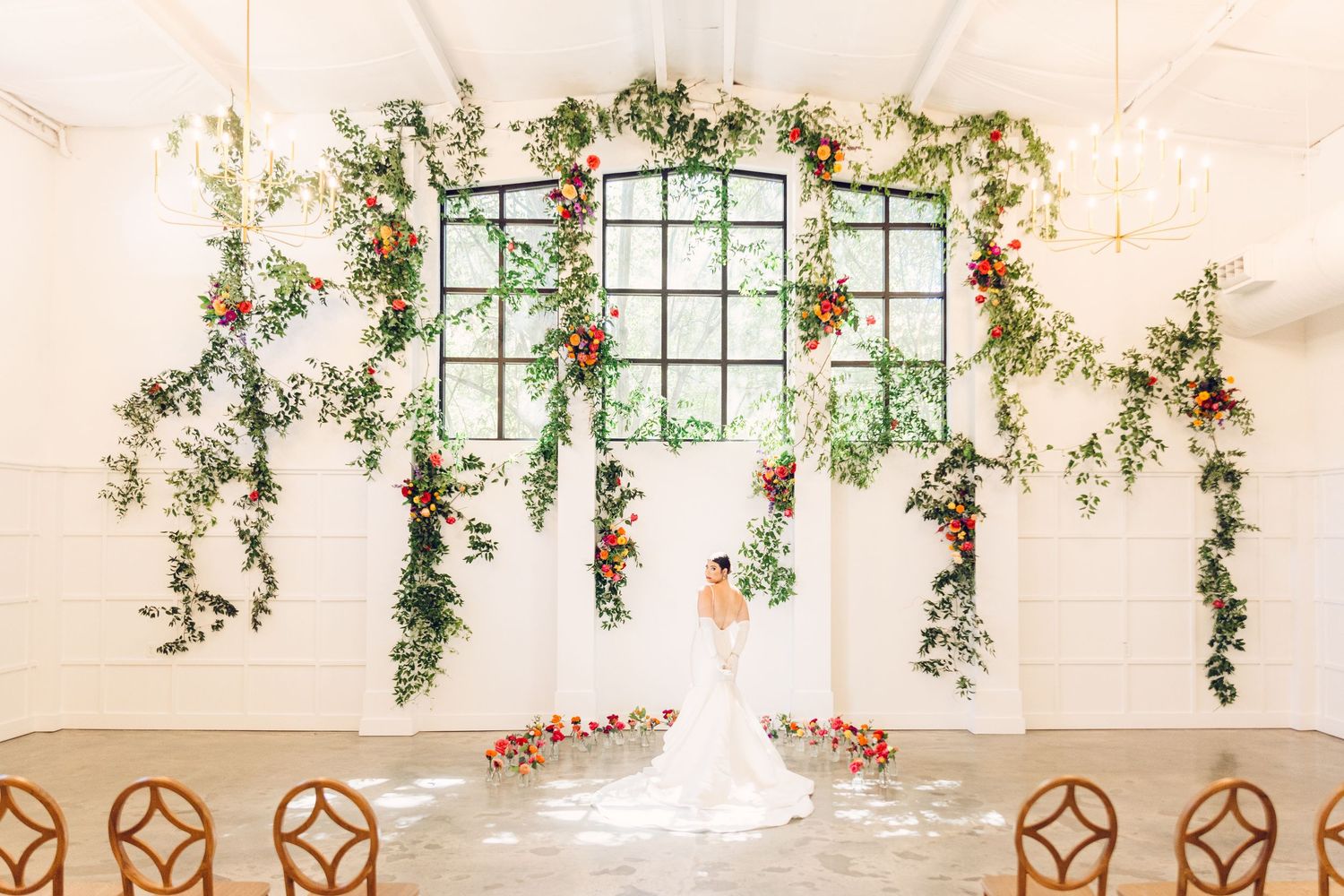 Elegant wedding reception venue with white draping, hanging greenery, and rustic wooden chairs arranged in rows.