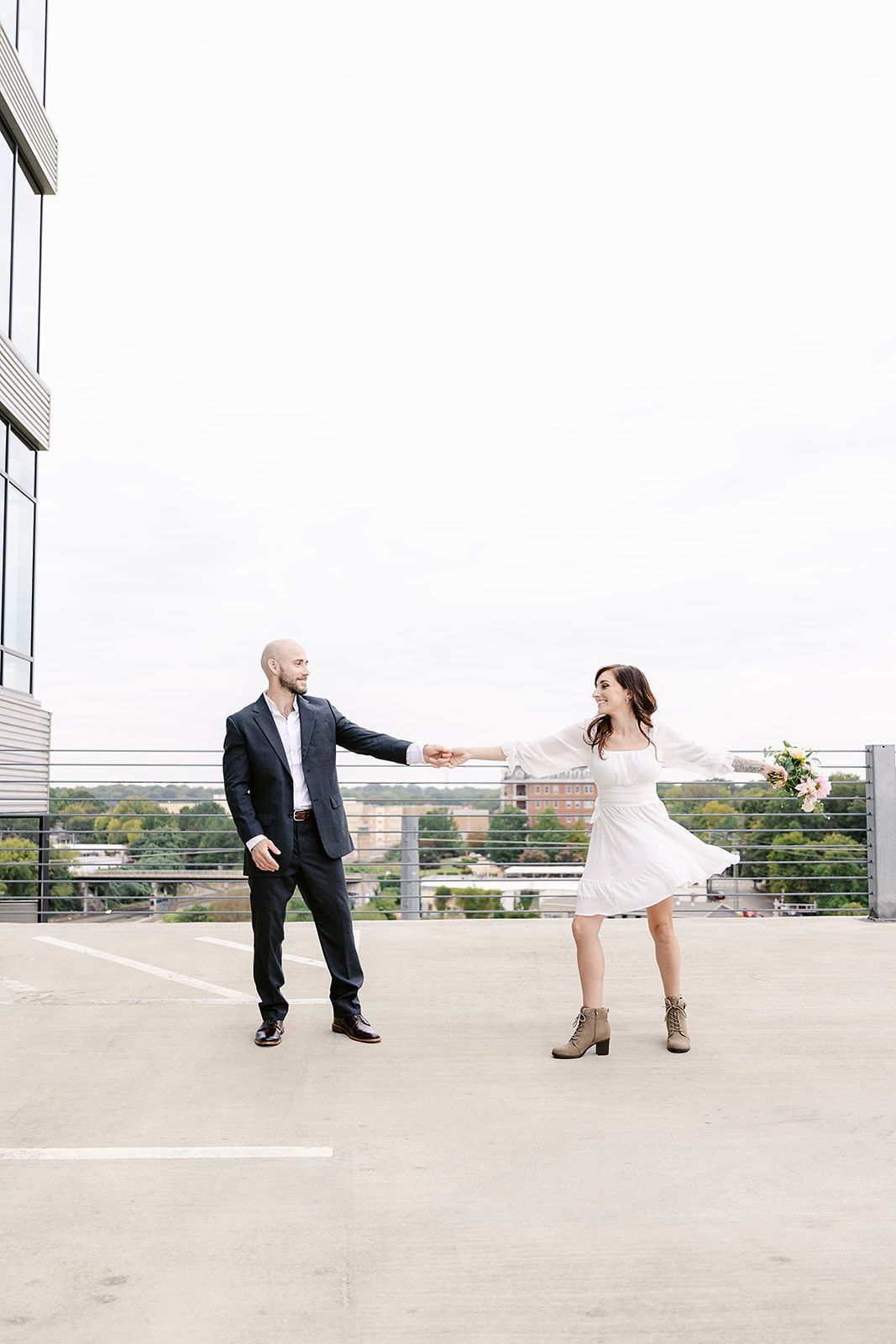 A couple dances together on a rooftop parking deck, with her in a white dress and cowboy boots and him in dark attire.