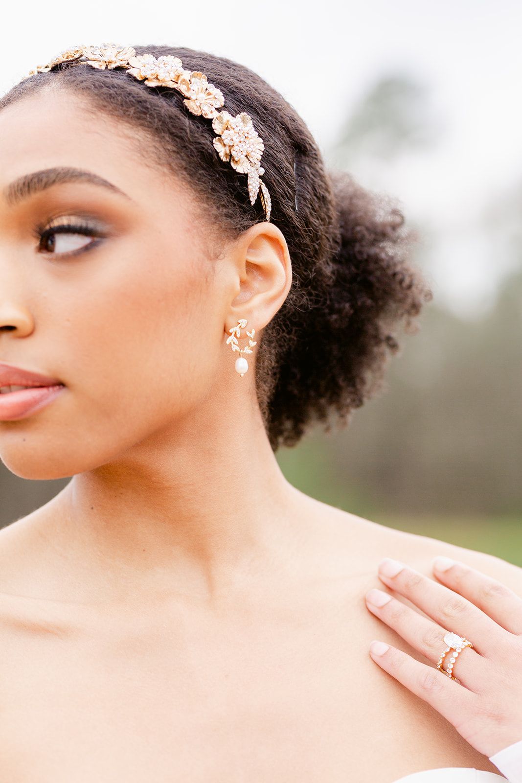 Close-up portrait showing delicate floral hairpiece, natural curls, and glowing makeup in soft outdoor lighting.