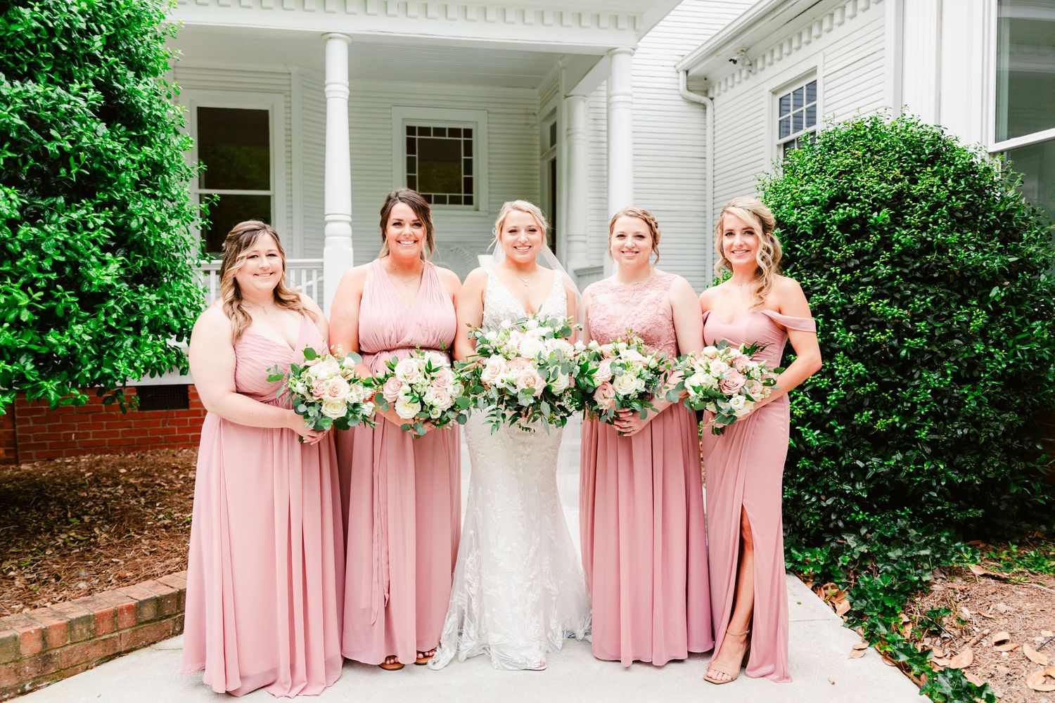 Bridal party in matching dusty rose dresses poses together in front of white building and green landscaping.