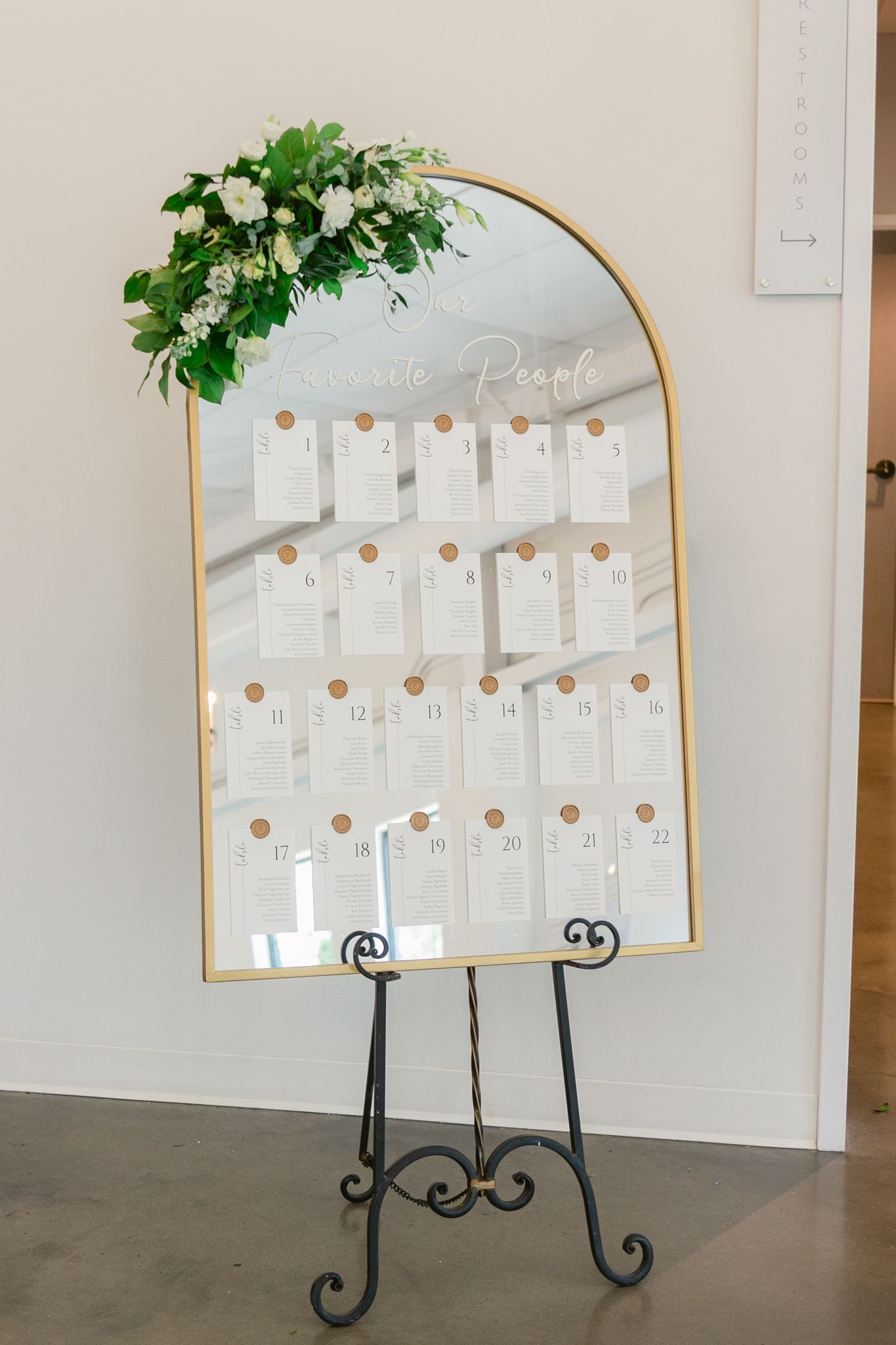 Elegant gold-framed seating chart display with greenery accents and white cards arranged in an arched design.