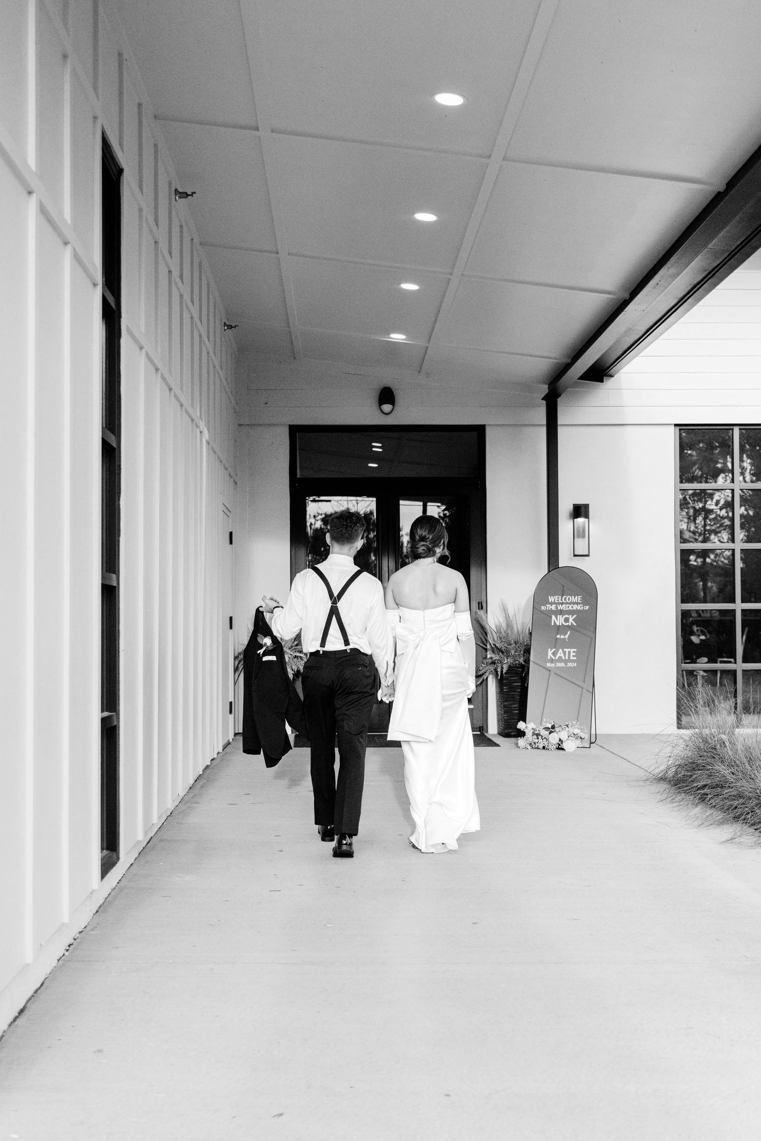 A black and white photo of a couple walking down a modern hallway toward glass doors.