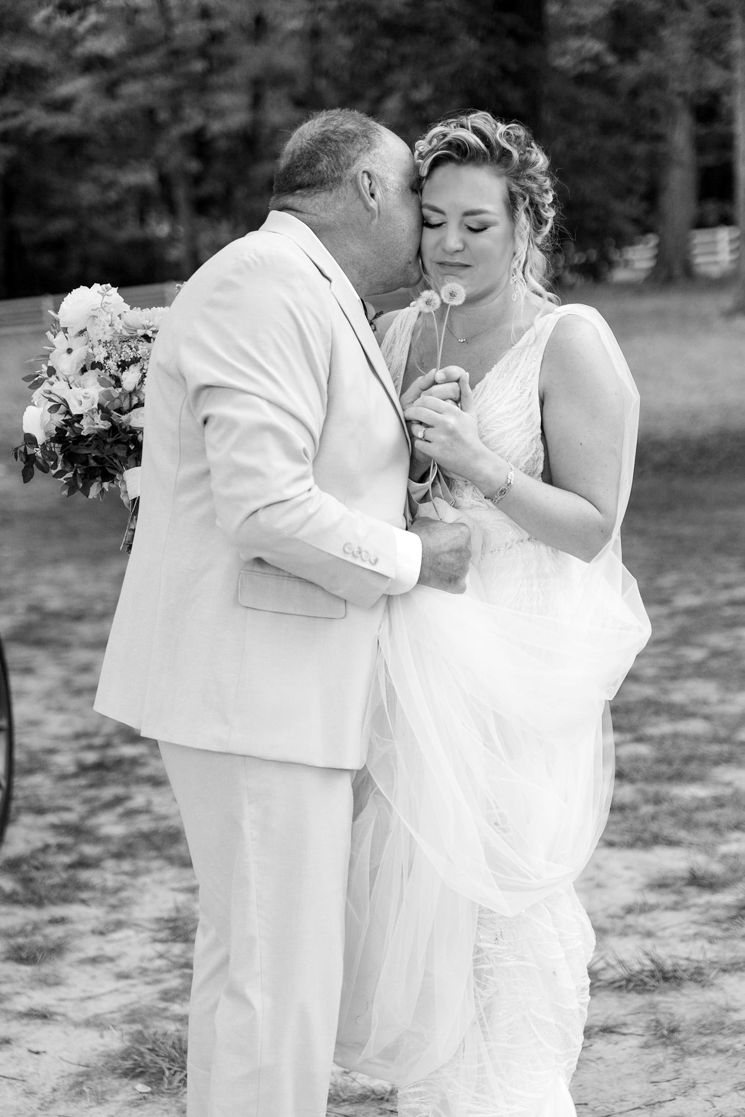 Black and white photo of a groom kissing bride on cheek while she holds a dandelion at outdoor wedding.