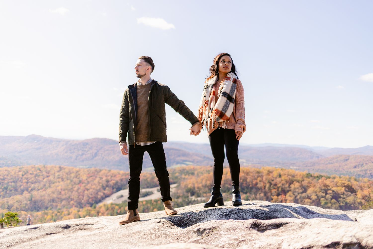 A couple stands together on a rocky mountain overlook with fall foliage and mountains visible in the distance.