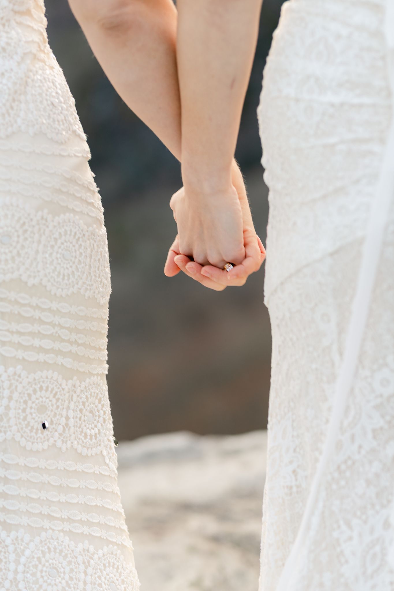 Close-up of two people in white lace wedding dresses holding hands against a blurred background.
