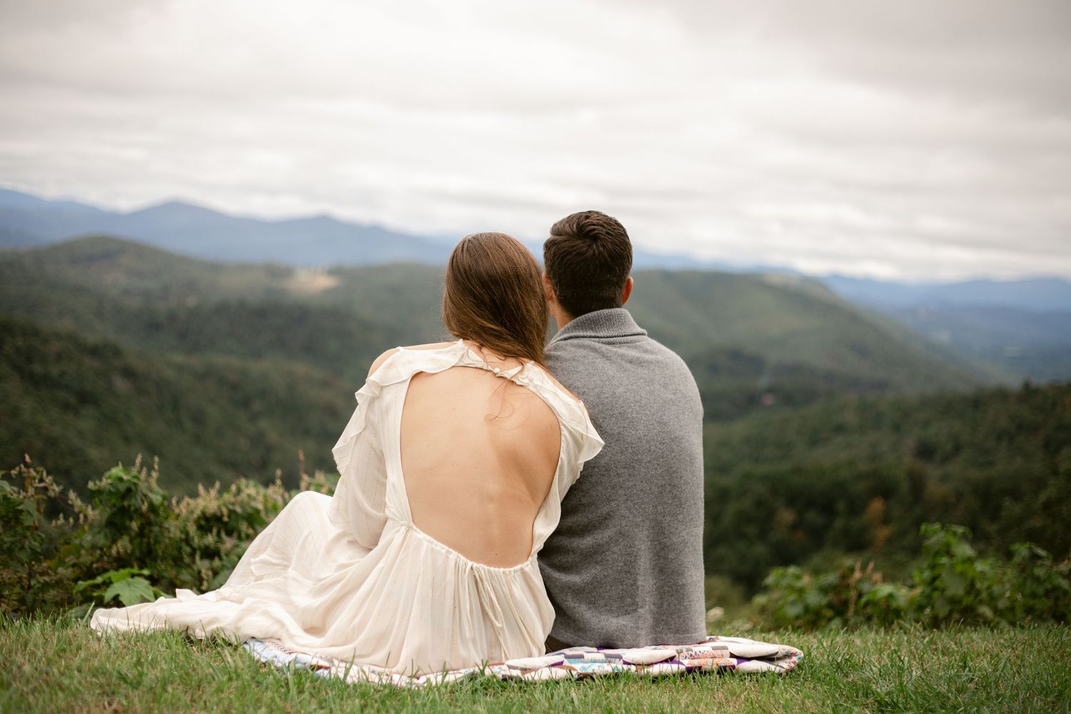 A couple sits on a blanket enjoying a scenic mountain vista on a cloudy day.