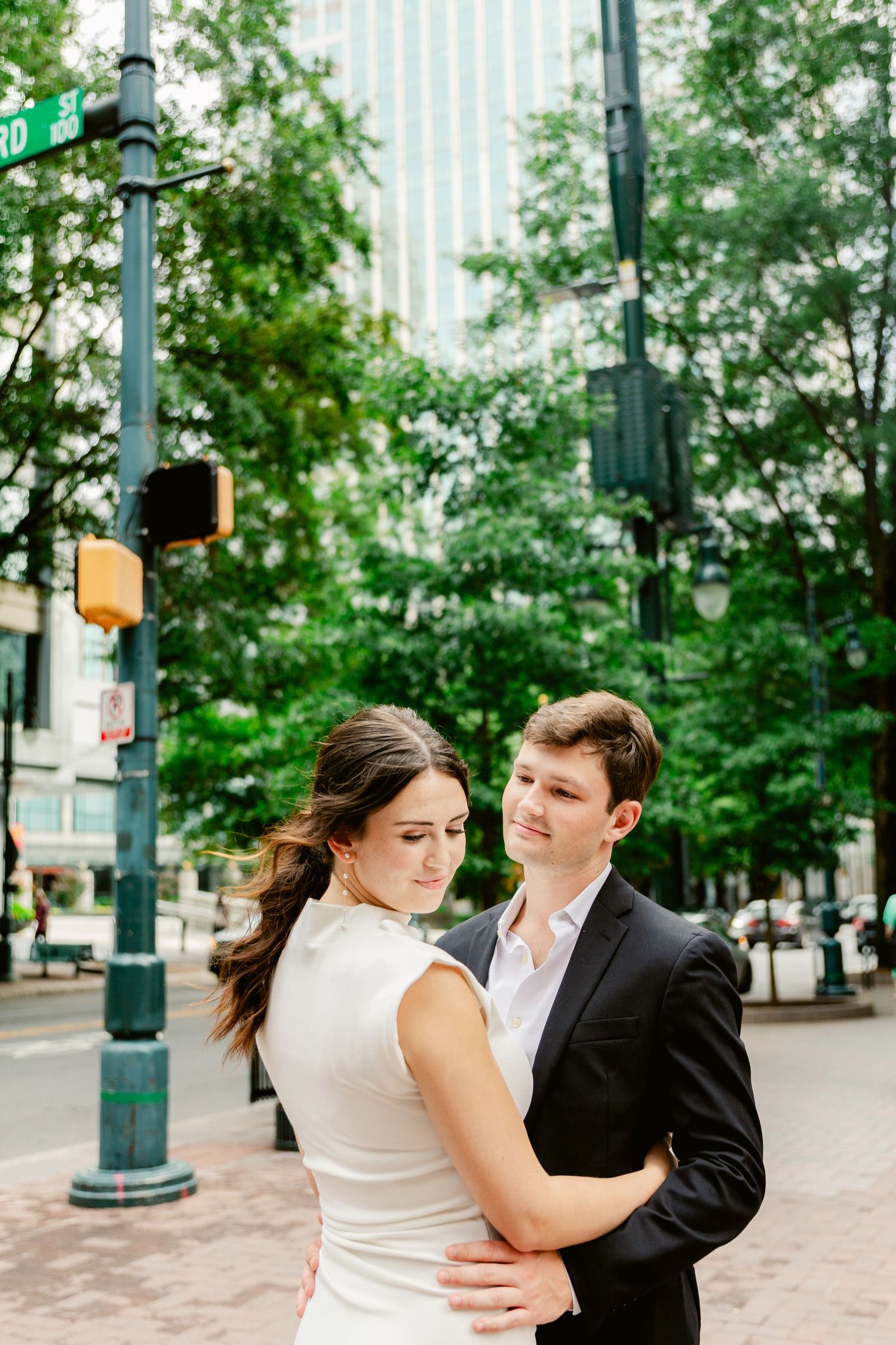 A couple embraces in a tender moment on a city street with lamp posts and trees in the background.