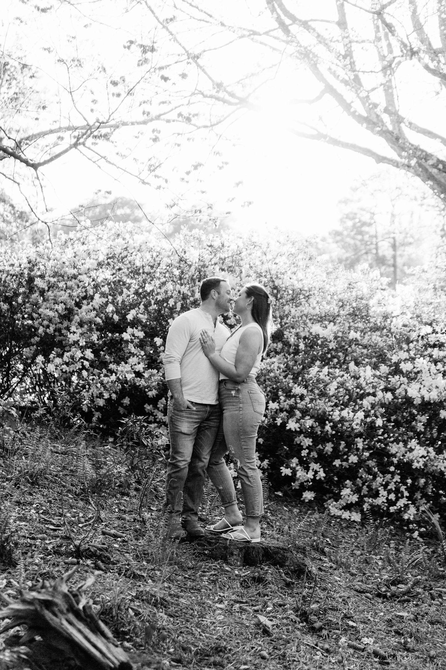 A black and white photo captures an intimate moment between a couple among flowering bushes in a garden setting.