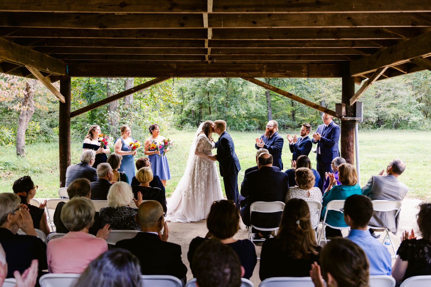 Outdoor wedding ceremony setup with white chairs and floral decorations under a rustic wooden pavilion.