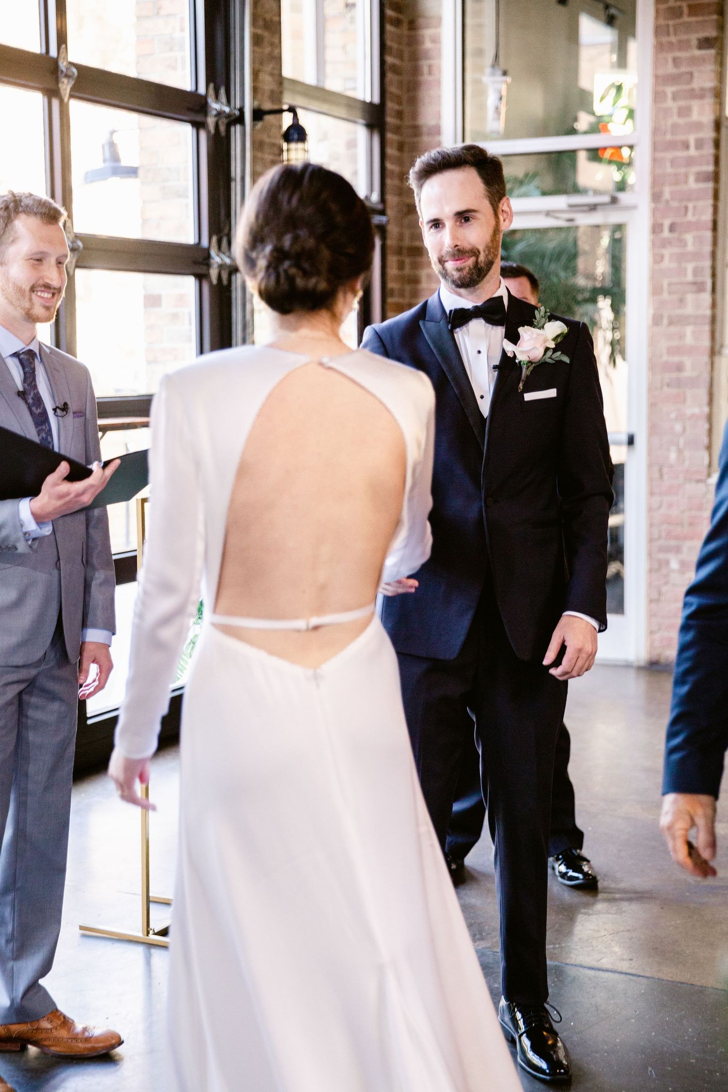 A wedding ceremony takes place in an industrial-chic venue with exposed brick walls and large windows.