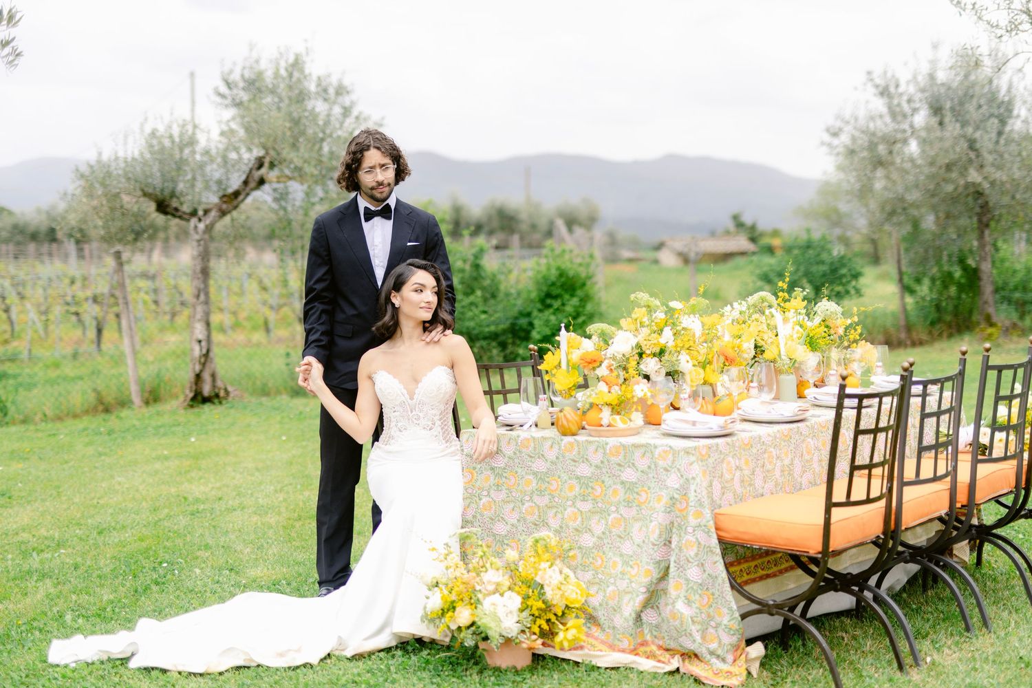 A rustic outdoor wedding reception table set with yellow floral centerpieces and wooden chairs in a scenic countryside setting.