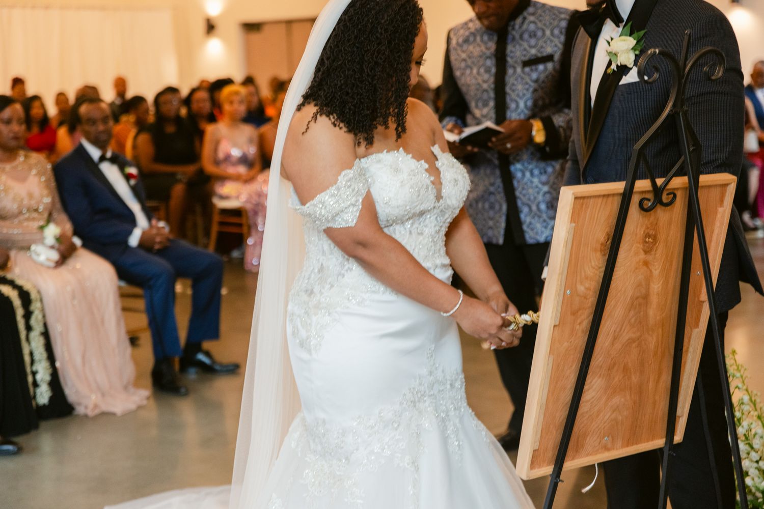 A wedding ceremony in progress with a white mermaid dress and veil against a wooden easel display.