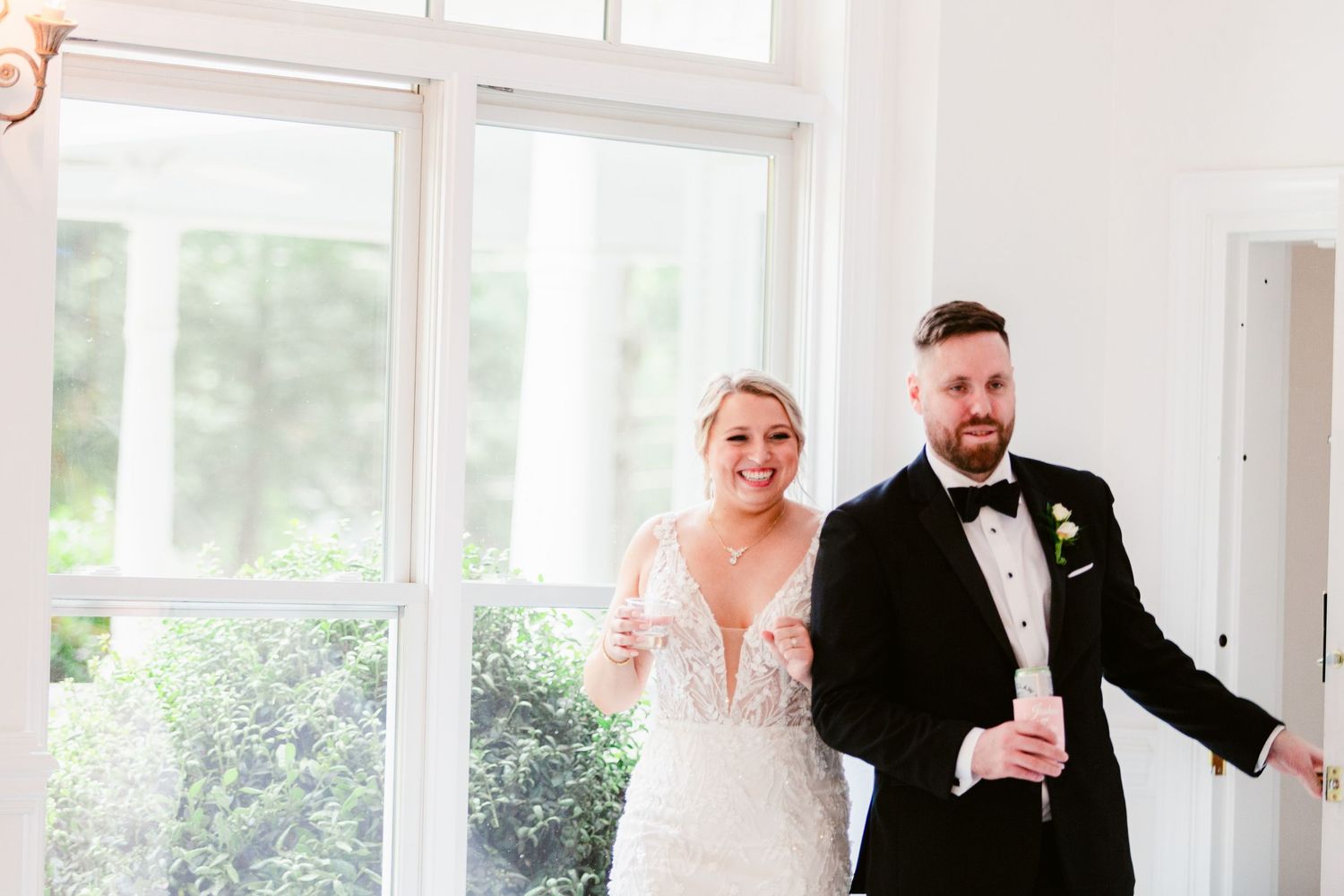 A wedding party stands together in a bright, sunlit room with white decor and large windows.
