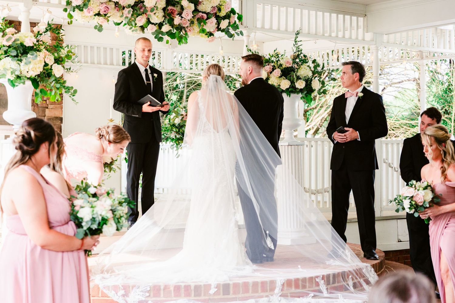 Wedding party gathers on a white porch decorated with pink and white floral arrangements for formal photographs.