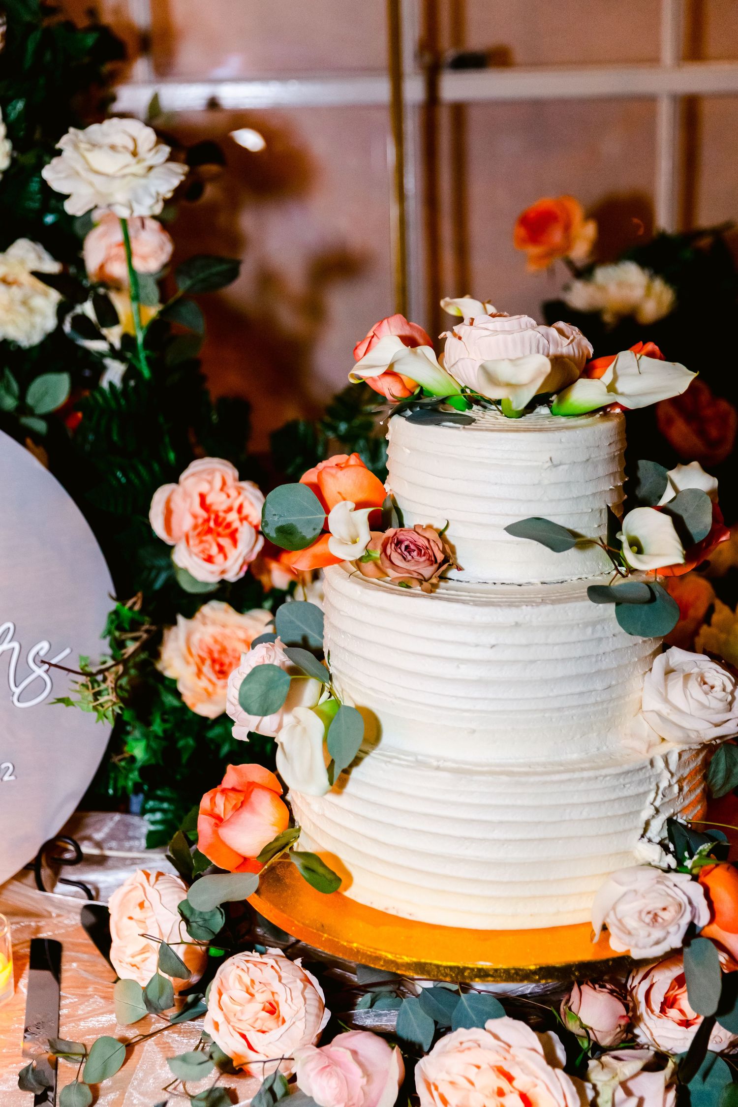Three-tiered white wedding cake decorated with peach roses and eucalyptus leaves on a wooden table.