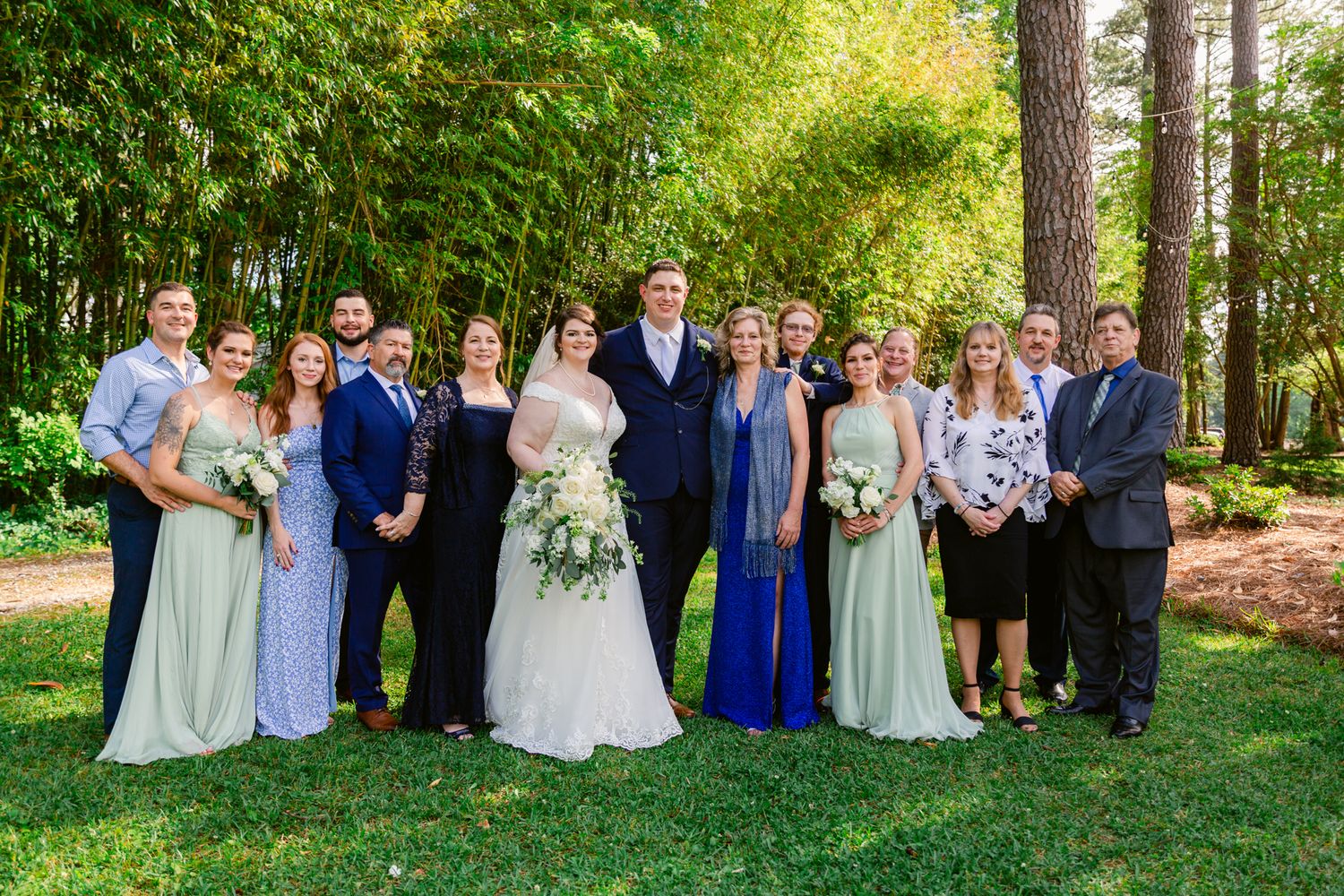 Wedding party poses together outdoors in a wooded setting with sunlight filtering through the trees.
