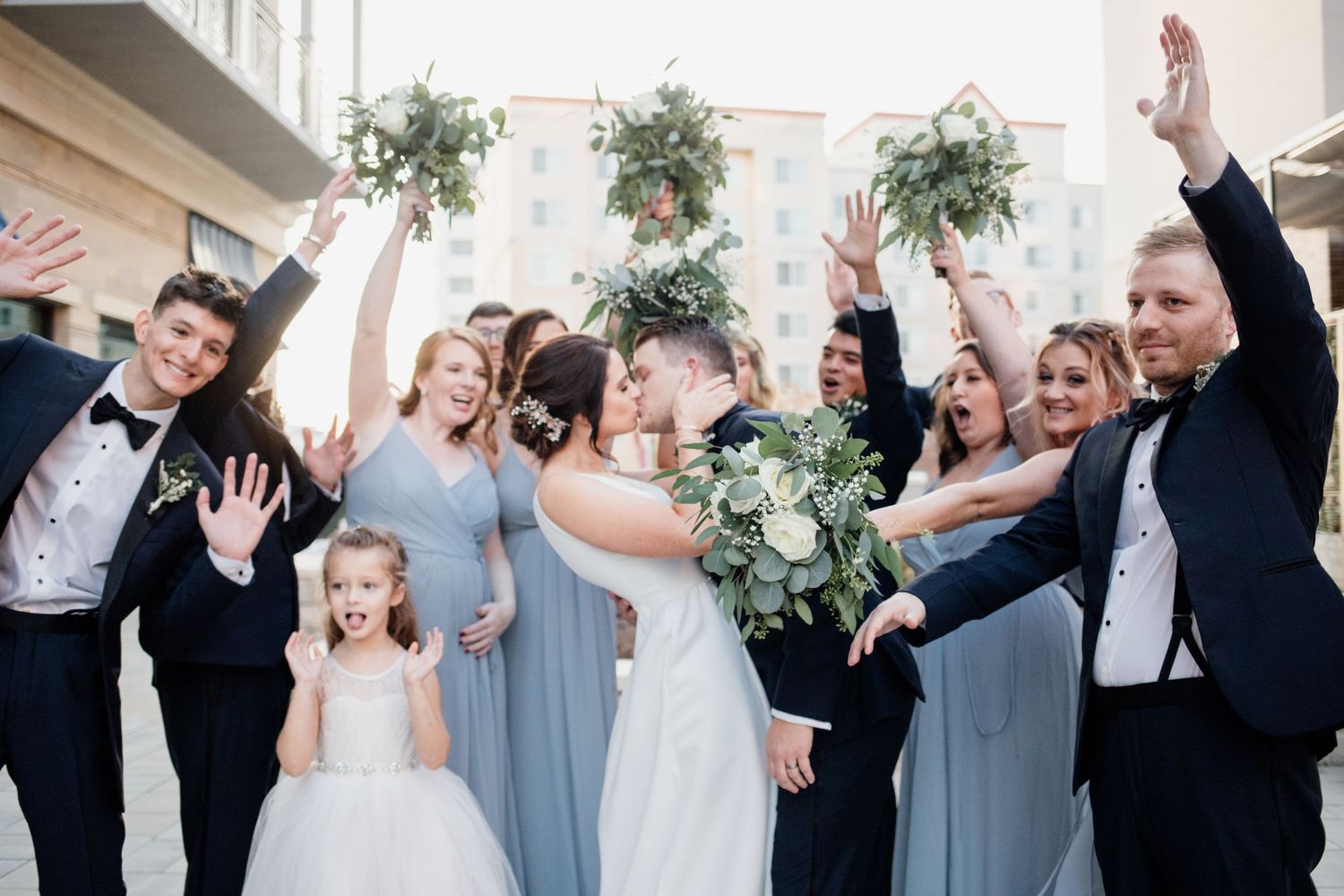 Wedding party celebrates with raised arms and floral arrangements in urban setting.