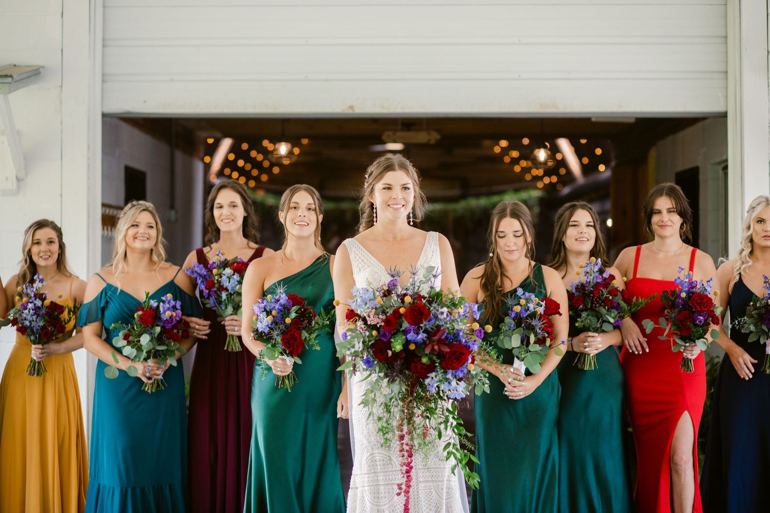 Wedding party lineup featuring bridesmaids in jewel-toned dresses holding colorful bouquets.