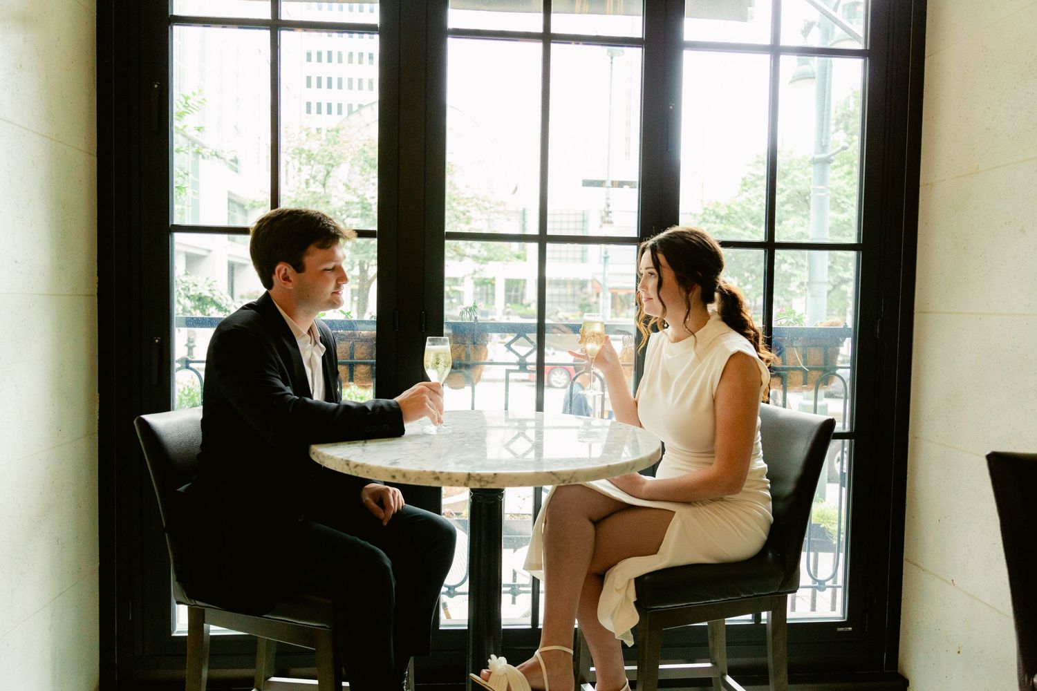 Two people having a professional conversation at a small cafe table near large windows in an elegant restaurant setting.