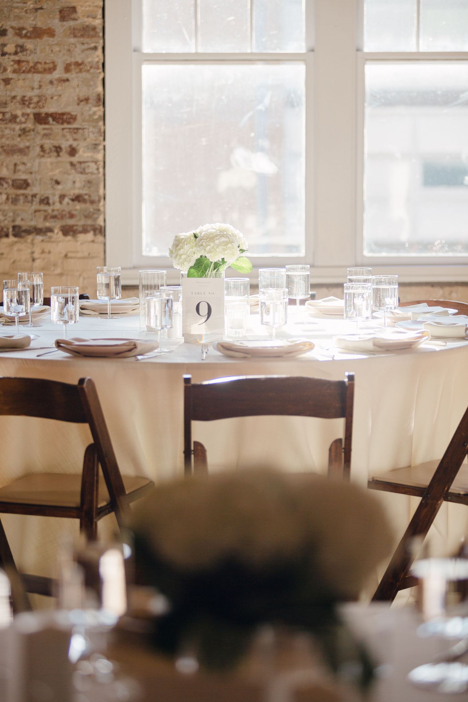 Round dining table with white tablecloth, place settings, and floral centerpiece near exposed brick wall.