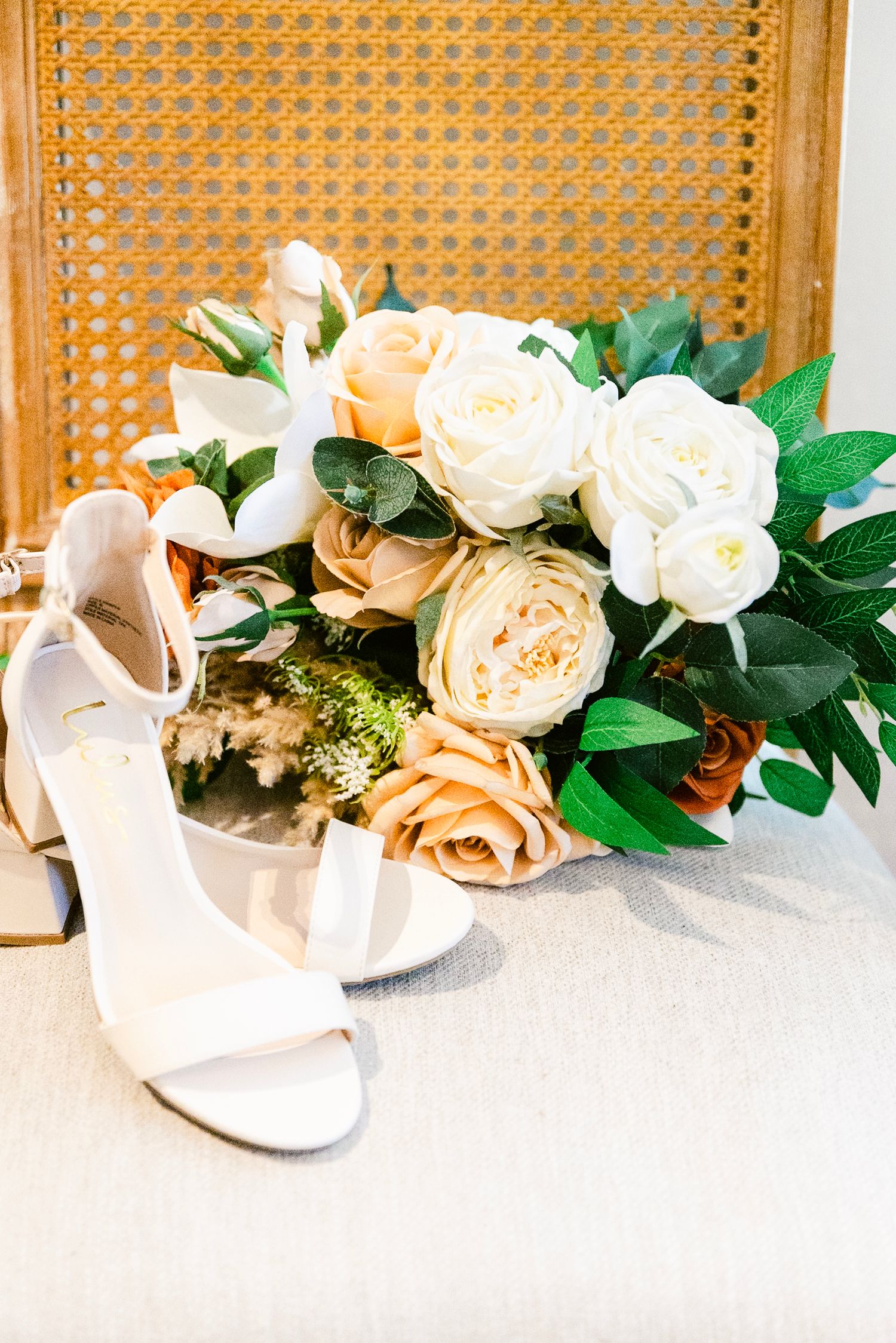 A beautiful bridal bouquet of peach and cream roses with greenery sits next to white wedding shoes.