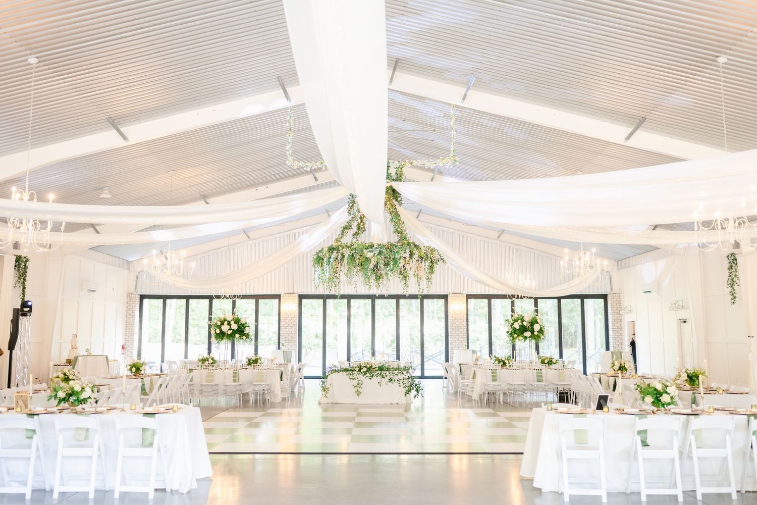 An elegant white wedding reception venue with high ceilings, large windows, and decorative greenery accents throughout.