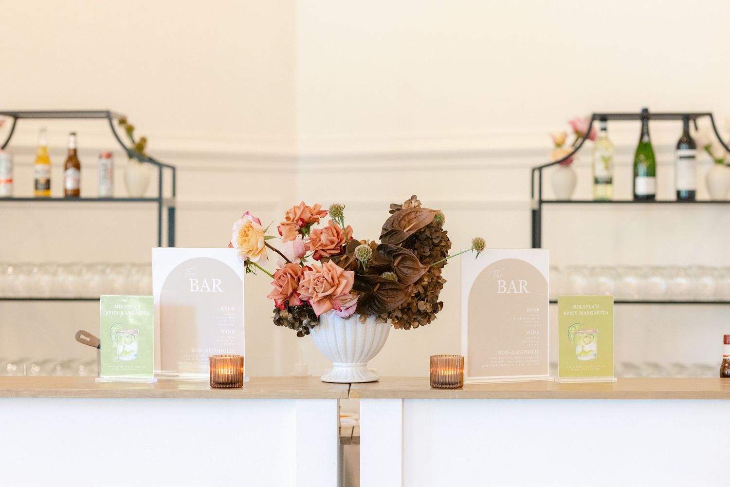 A soft pink and white event space decorated with elegant floral arrangements and candles on wooden tables.