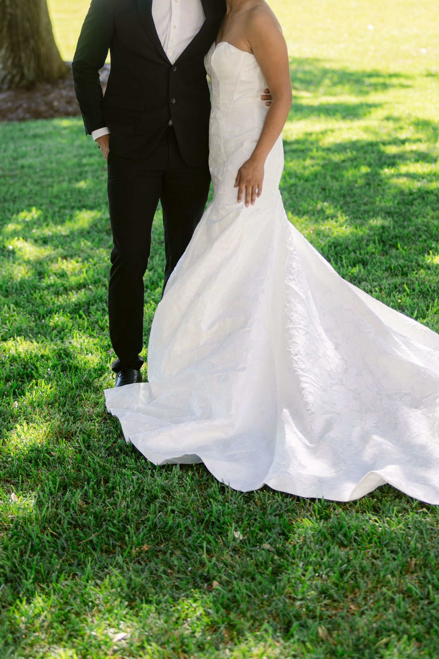 A stunning white wedding dress with a flowing train contrasts against lush green grass at an outdoor wedding.