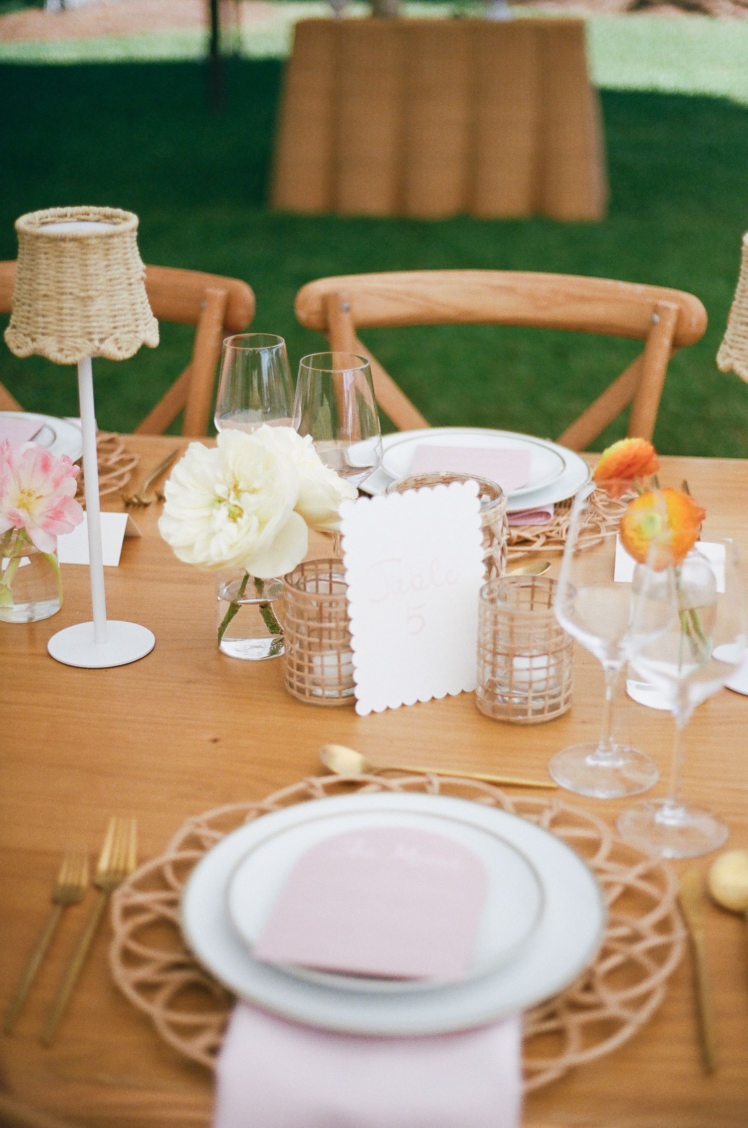 Elegant table setting with white plates, glassware, and delicate floral centerpieces on a wooden table for an outdoor event.
