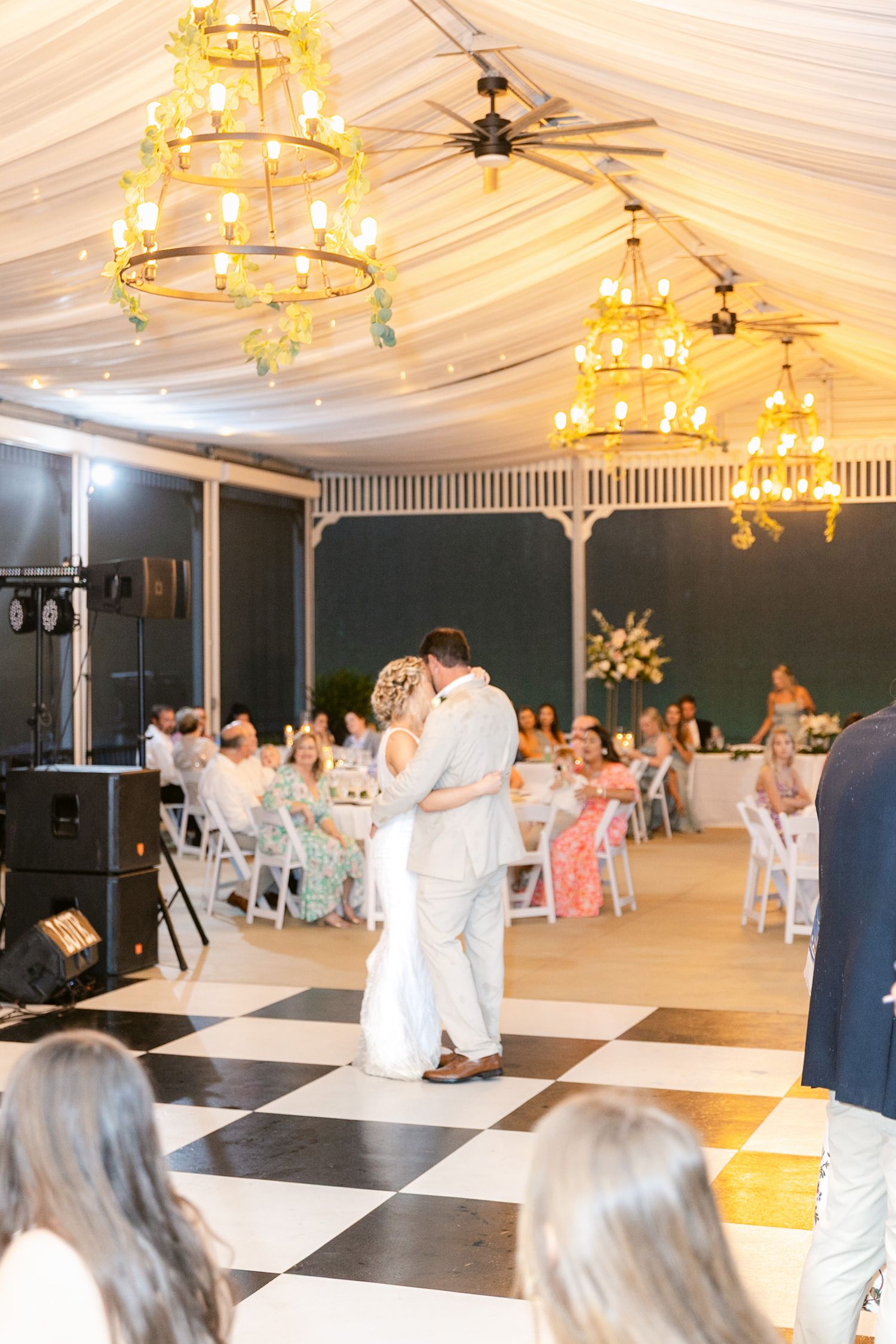 A wedding reception venue with a black and white checkered dance floor under elegant gold chandeliers in a white tent.