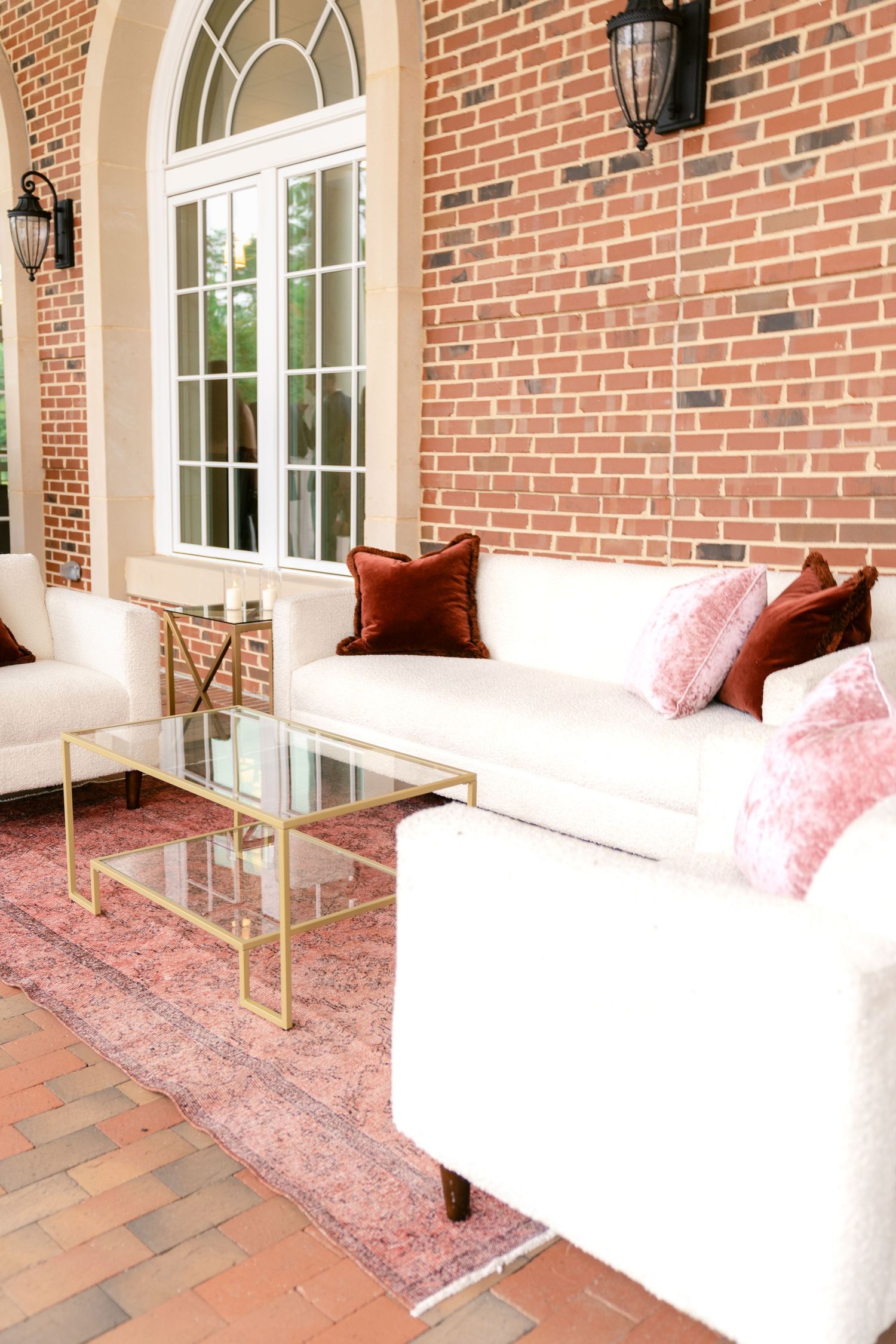 Elegant sunroom with white furniture, brick walls, and a pink vintage rug features glass doors and classic lantern sconces.