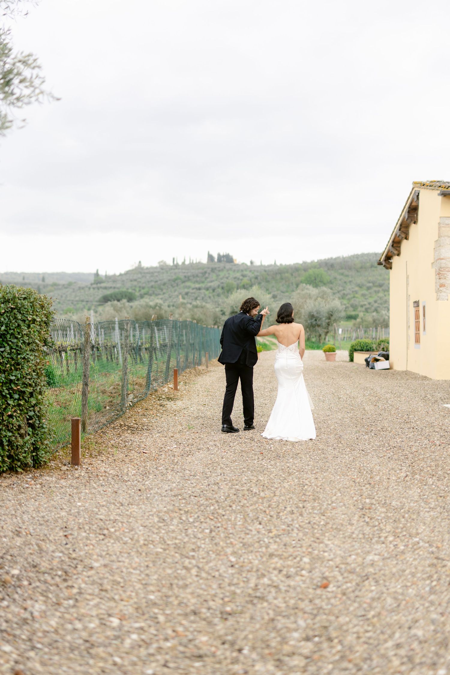 A couple in formal attire walks along a gravel path beside a vineyard in the Tuscan countryside.