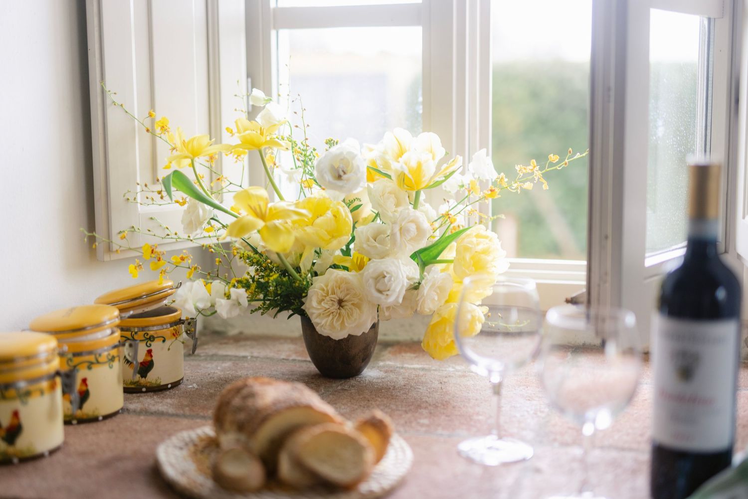 A bright spring tablescape with yellow flowers, white roses, and wine bottles arranged near sunlit windows.