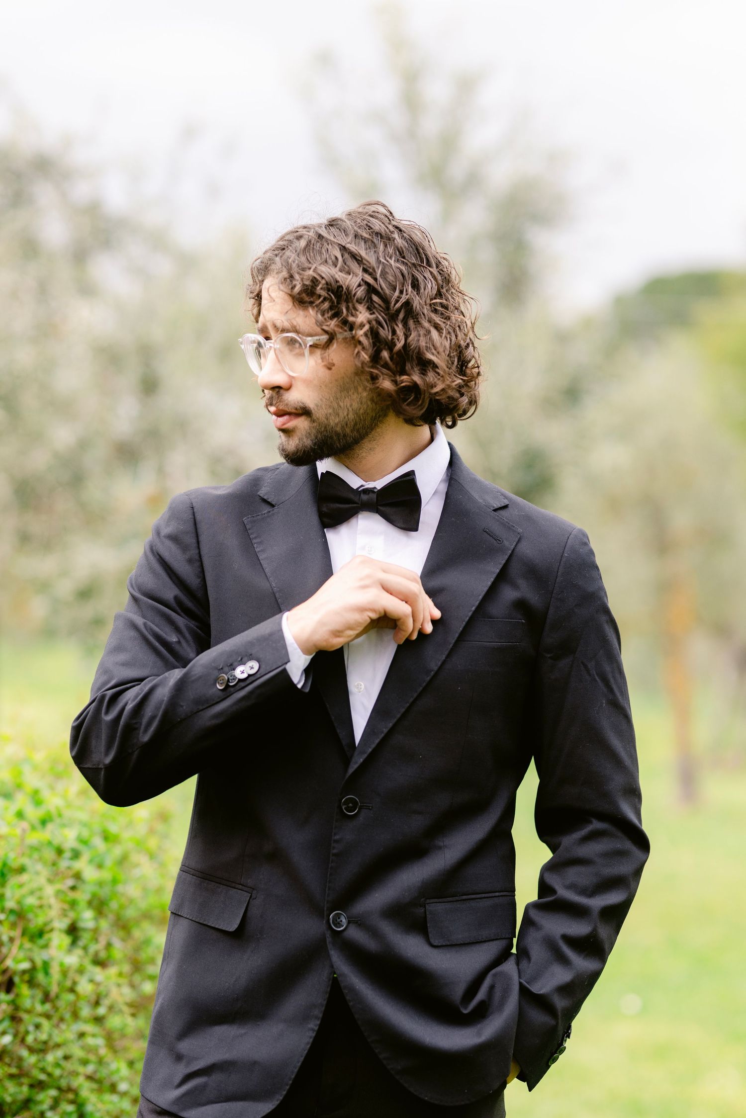 A person in a black tuxedo and bowtie poses thoughtfully in an outdoor garden setting.