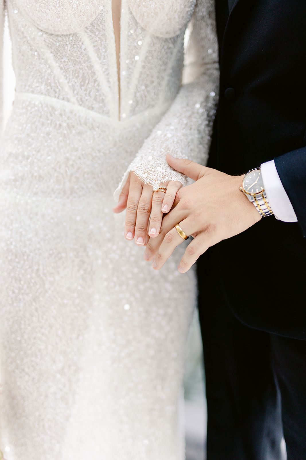 Close-up of newlywed couple's hands showing wedding rings and bride's sparkly white lace dress sleeve.