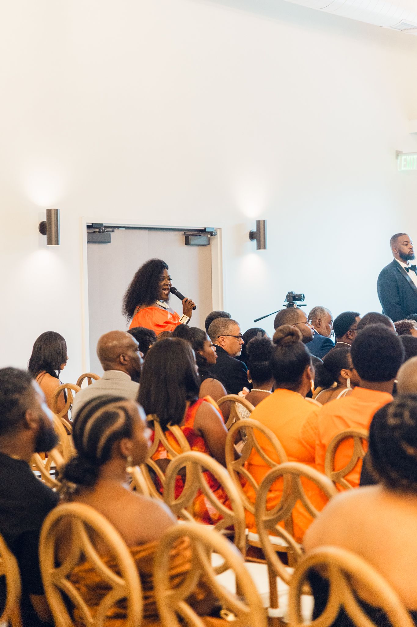 Attendees in bright orange outfits seated in rows at an indoor ceremony.