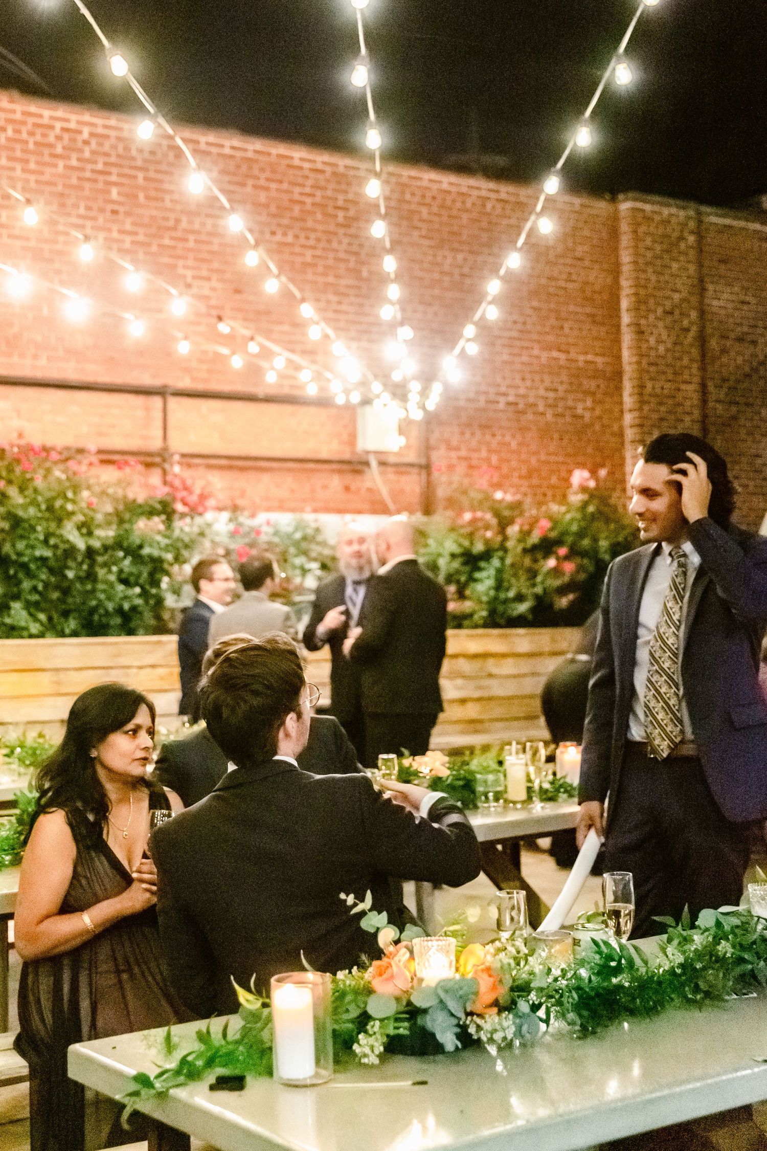 People gather at an outdoor evening event under string lights with candles and greenery decorating long tables.