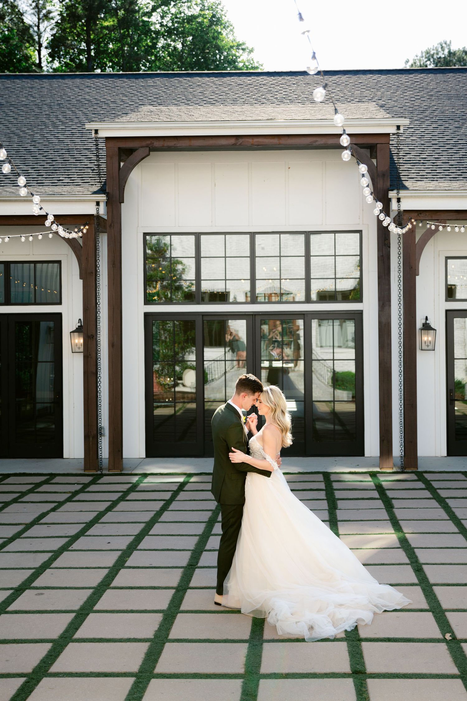 A romantic wedding couple shares an intimate moment on a patterned patio beneath string lights and elegant architecture.