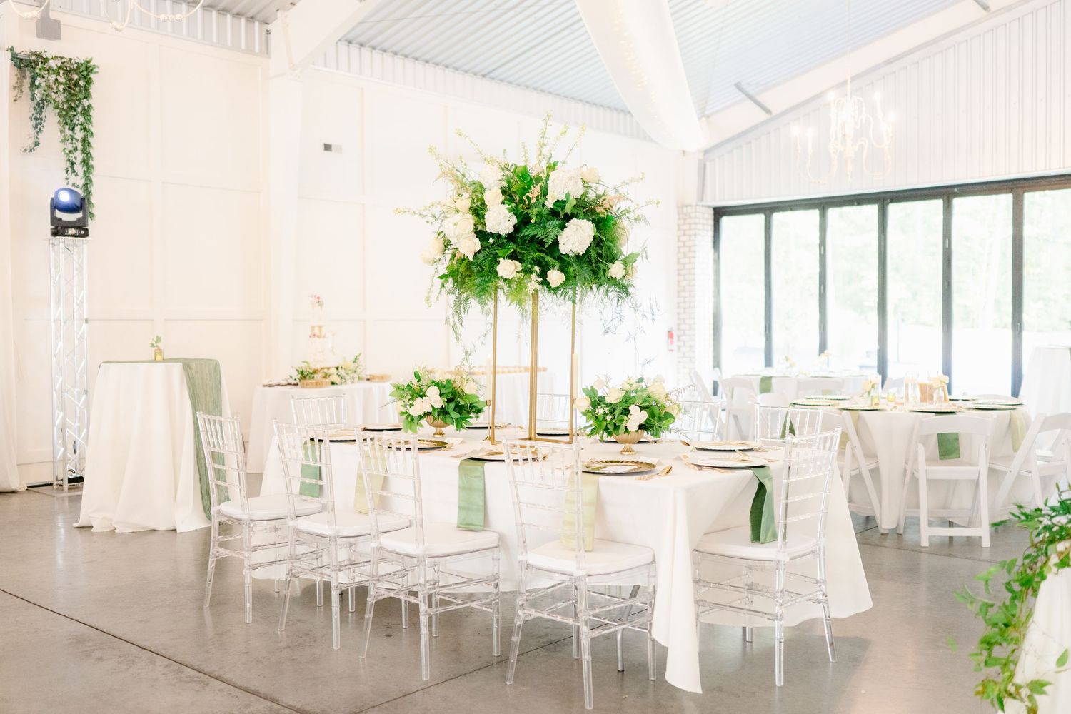 Elegant white wedding venue interior with round tables, floral centerpieces, and natural light streaming through windows.