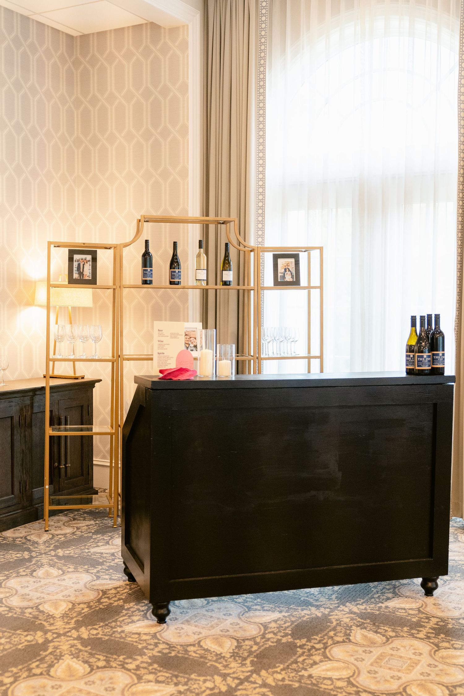 Elegant black mobile bar cart with gold shelving unit and wine bottles displayed against beige wallpapered wall.