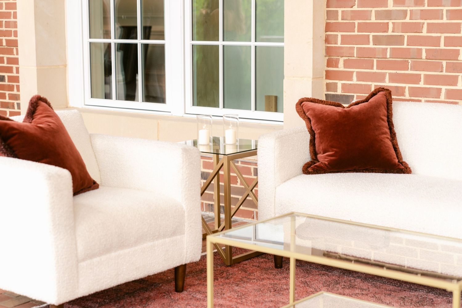 Bright brick-walled patio room with white upholstered seating, burgundy pillows, and brass accent tables on a pink area rug.