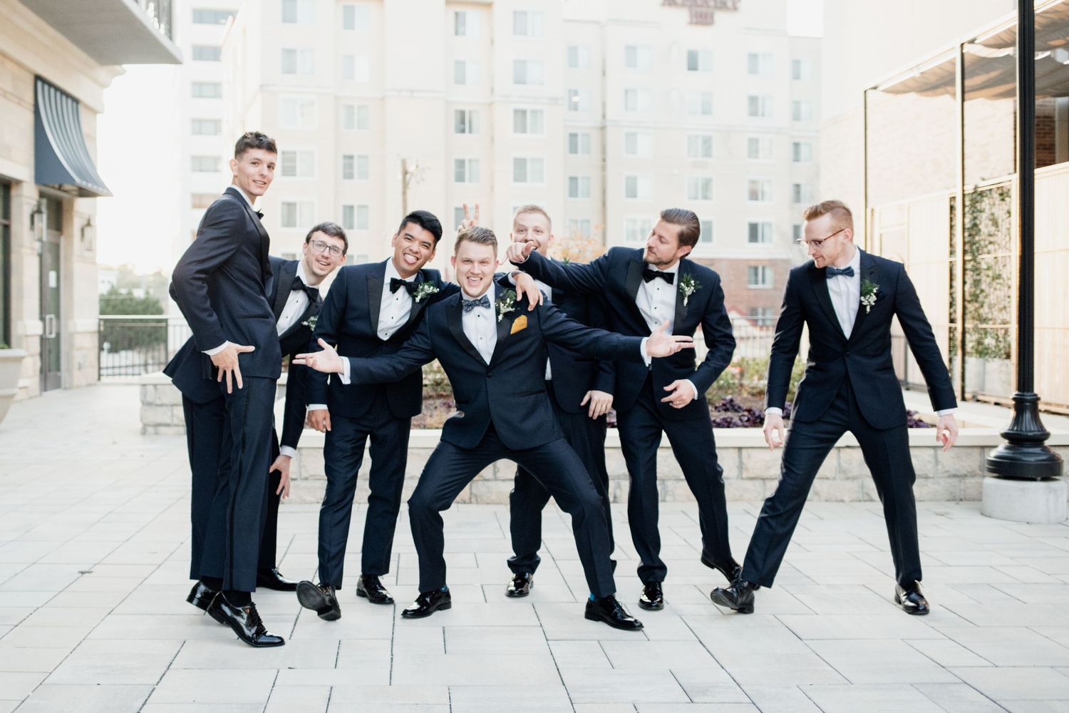 Groomsmen in black tuxedos strike playful poses together on a paved outdoor walkway.