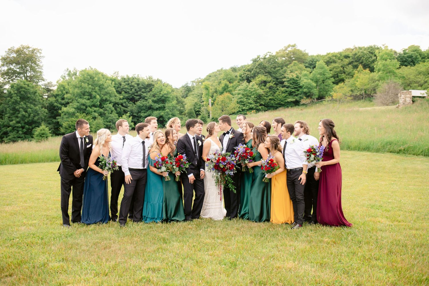 Wedding party group photo in field with bridesmaids in mixed jewel-toned dresses.