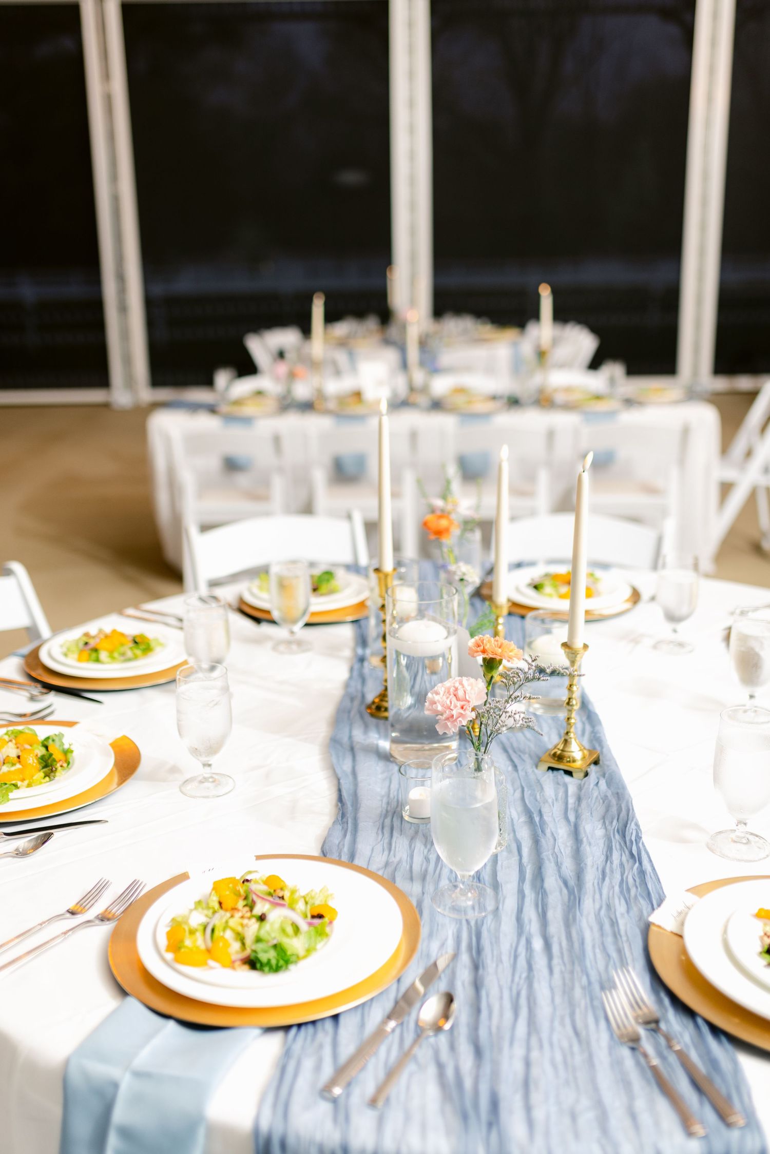 Elegant wedding table setup with light blue chiffon runner, white plates, and gold accents in natural lighting.