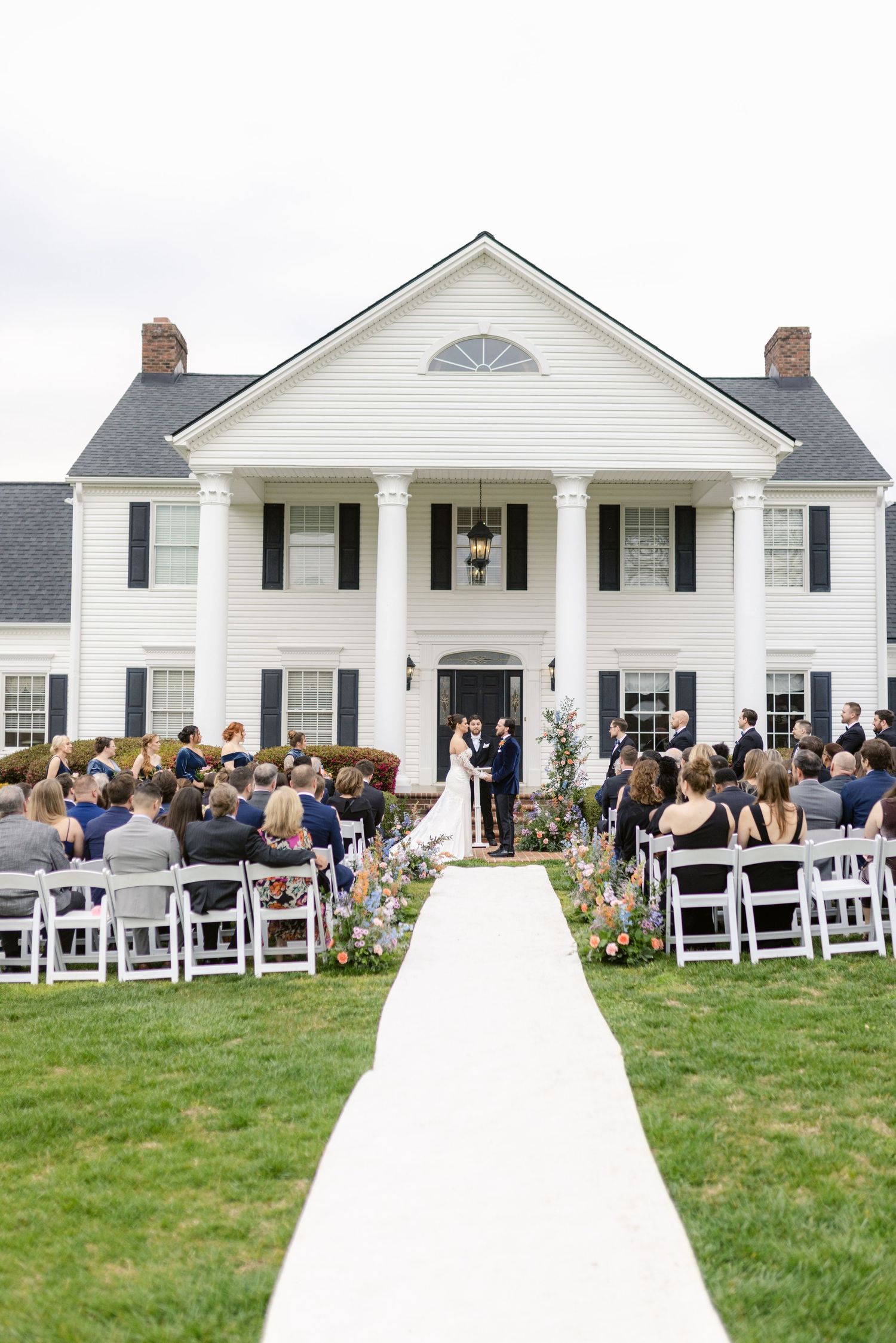Elegant outdoor wedding ceremony with white chairs arranged in front of a stately white colonial mansion with black shutters.