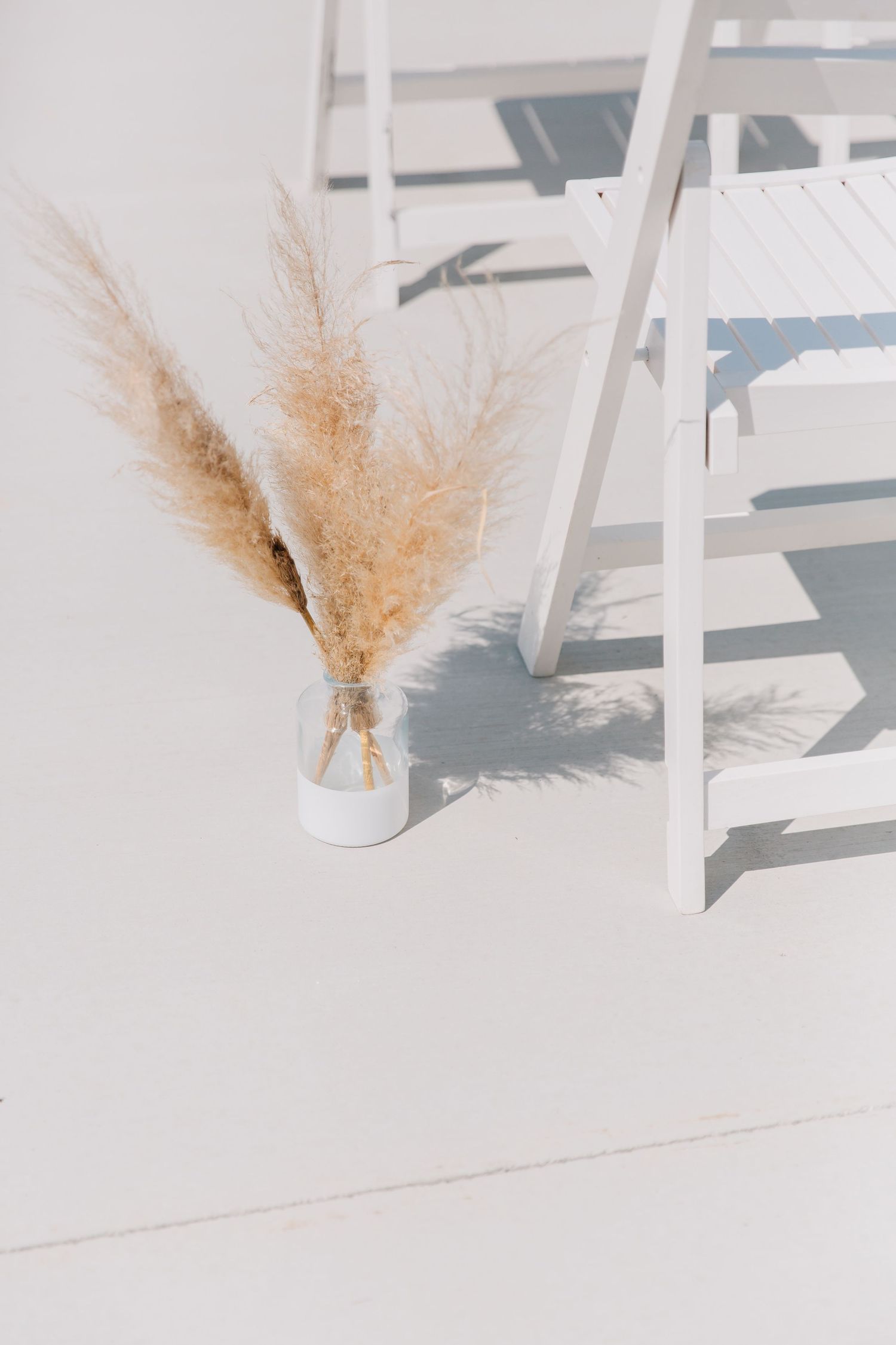 Dried pampas grass in white vase beside white ceremony chairs.